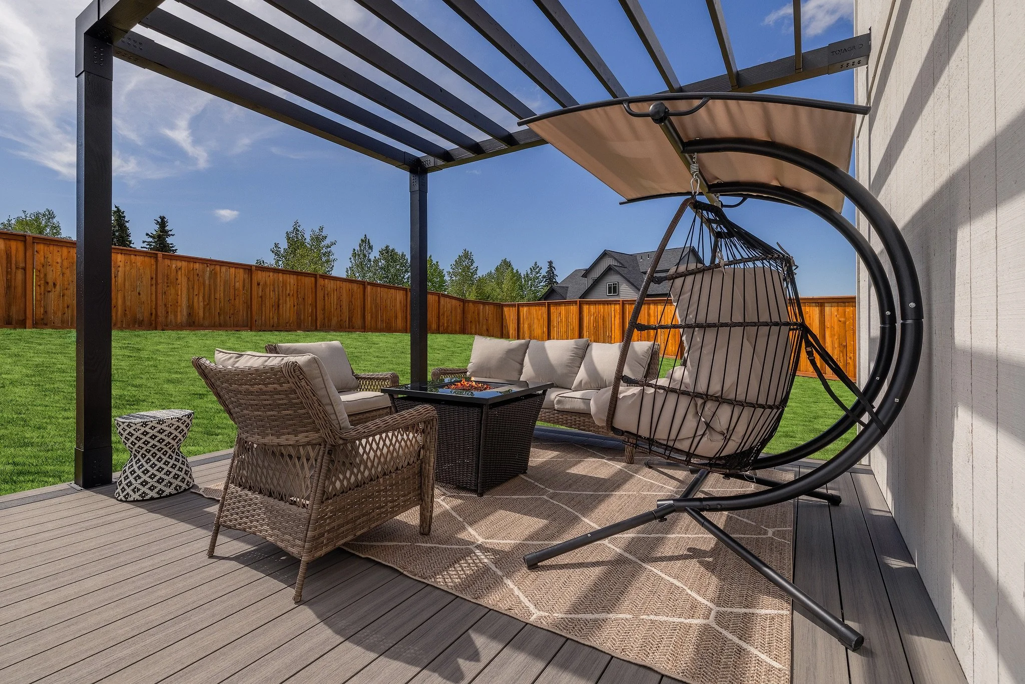 Outdoor patio with wicker chairs, a beige couch, a hanging swing chair, a fire pit, and a small table on a composite deck, enclosed by a wooden fence with a house and trees in the background under a cloudy sky.