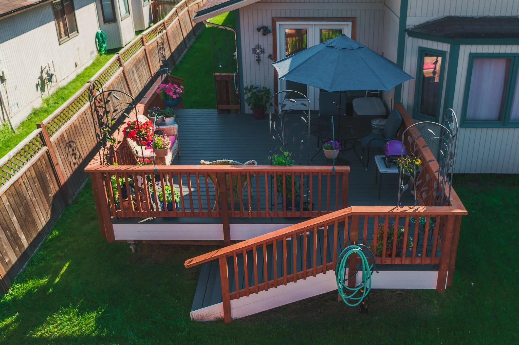 A backyard trex deck with outdoor furniture, potted plants, a garden hose, and an umbrella, surrounded by a grassy yard and fence.