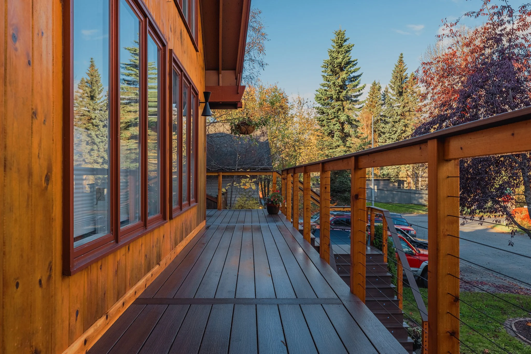 Composite decking balcony with cedar railing, potted plant, view of trees and parked cars, part of a house with wooden siding, clear blue sky.