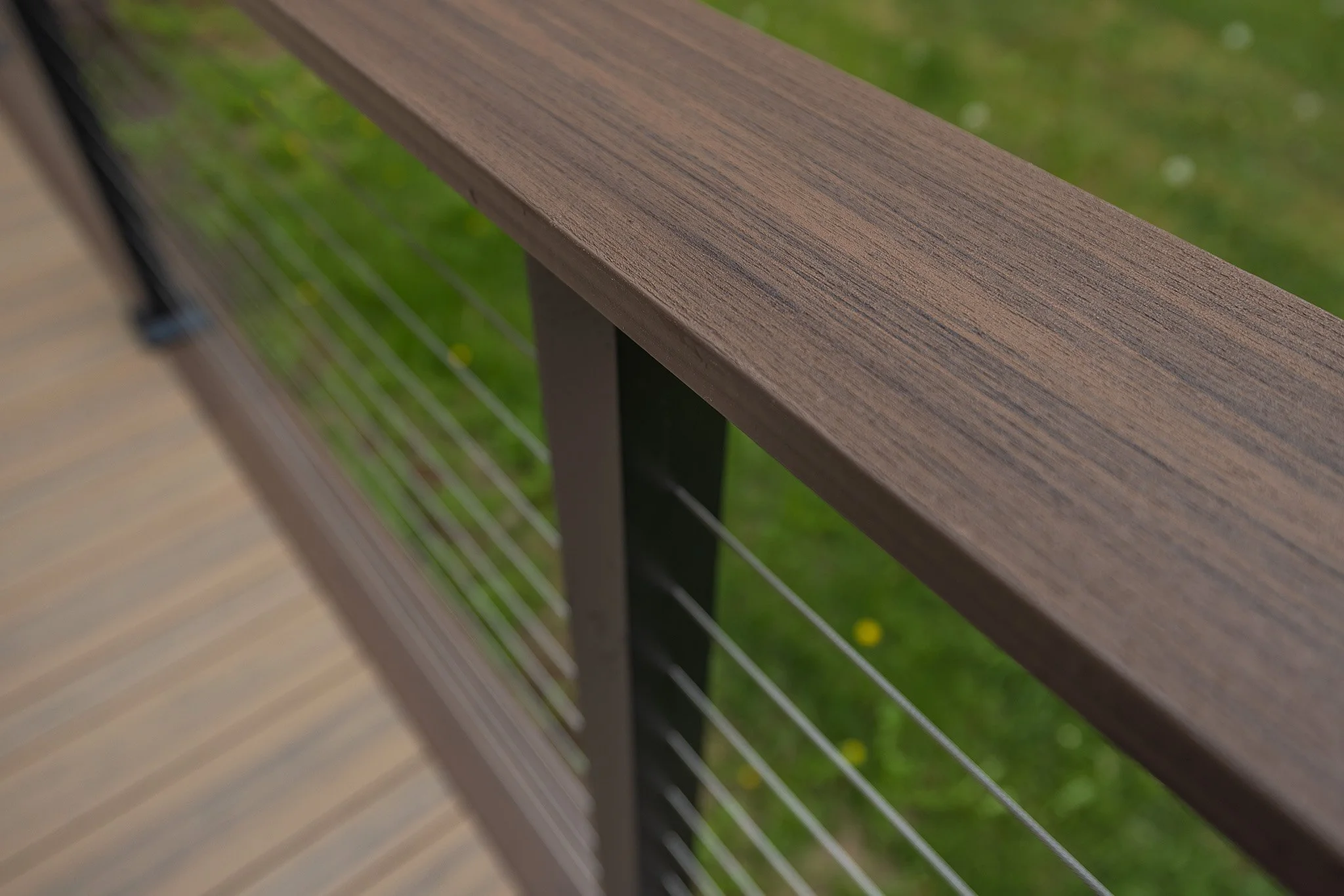 Close-up of a aluminum cable railing with a composite top cap, with green grass and small yellow flowers visible in the background.
