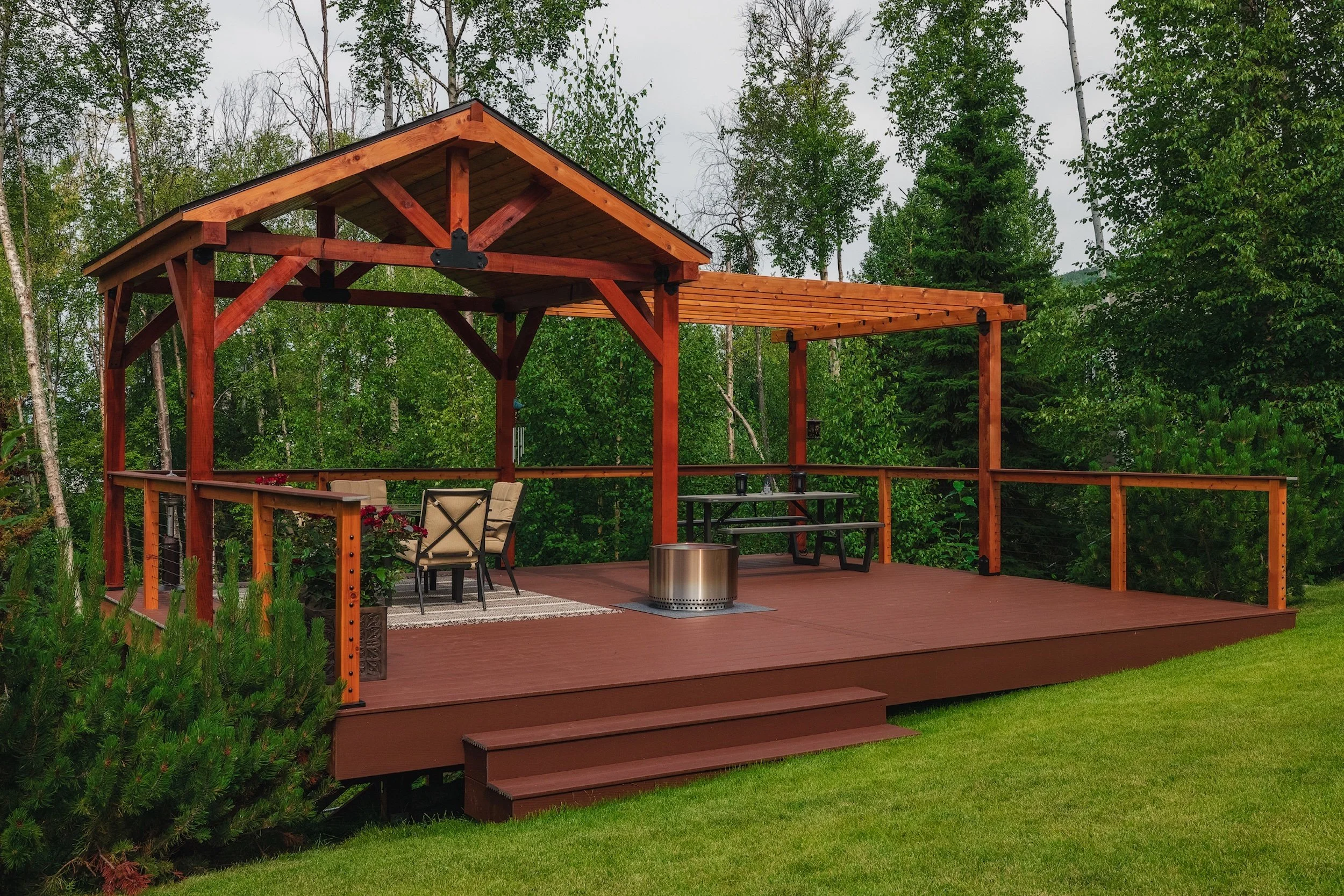 A wcomposite outdoor deck with steps leading up to it, surrounded by green grass and trees. The deck has a covered section with chairs, a table, a barbecue grill, and decorative plants.