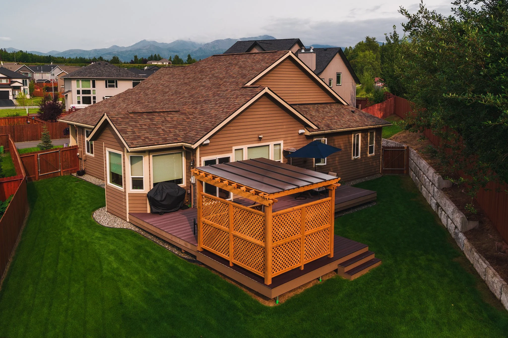 A backyard with a trex deck featuring a lattice privacy screen and a covered patio area with an umbrella, lush green grass, and a fenced perimeter. There are neighboring houses and a mountain range in the background.