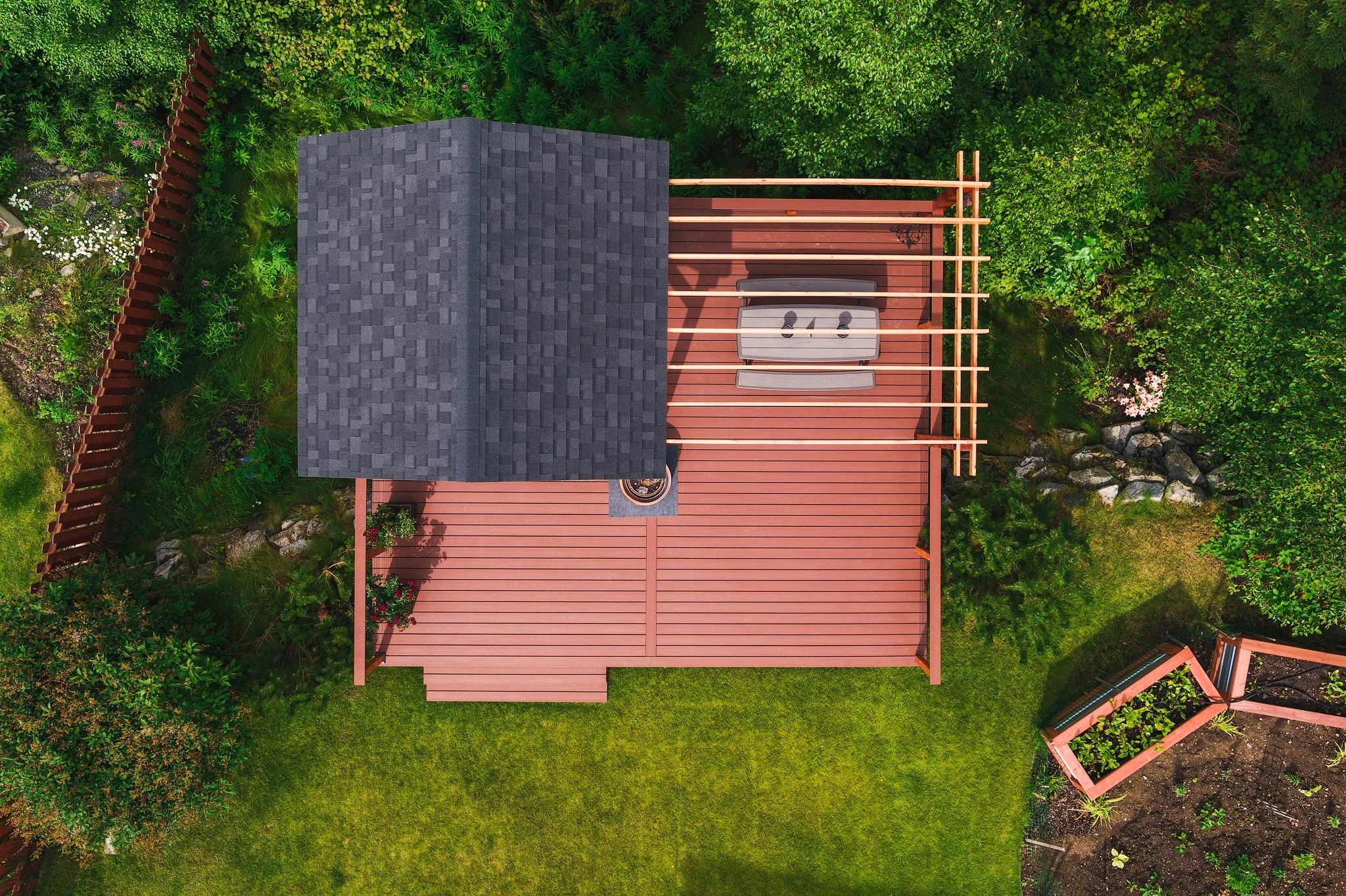Aerial view of a backyard trex deck with a small shelter, a bench, and a fire pit, surrounded by green trees and bushes.