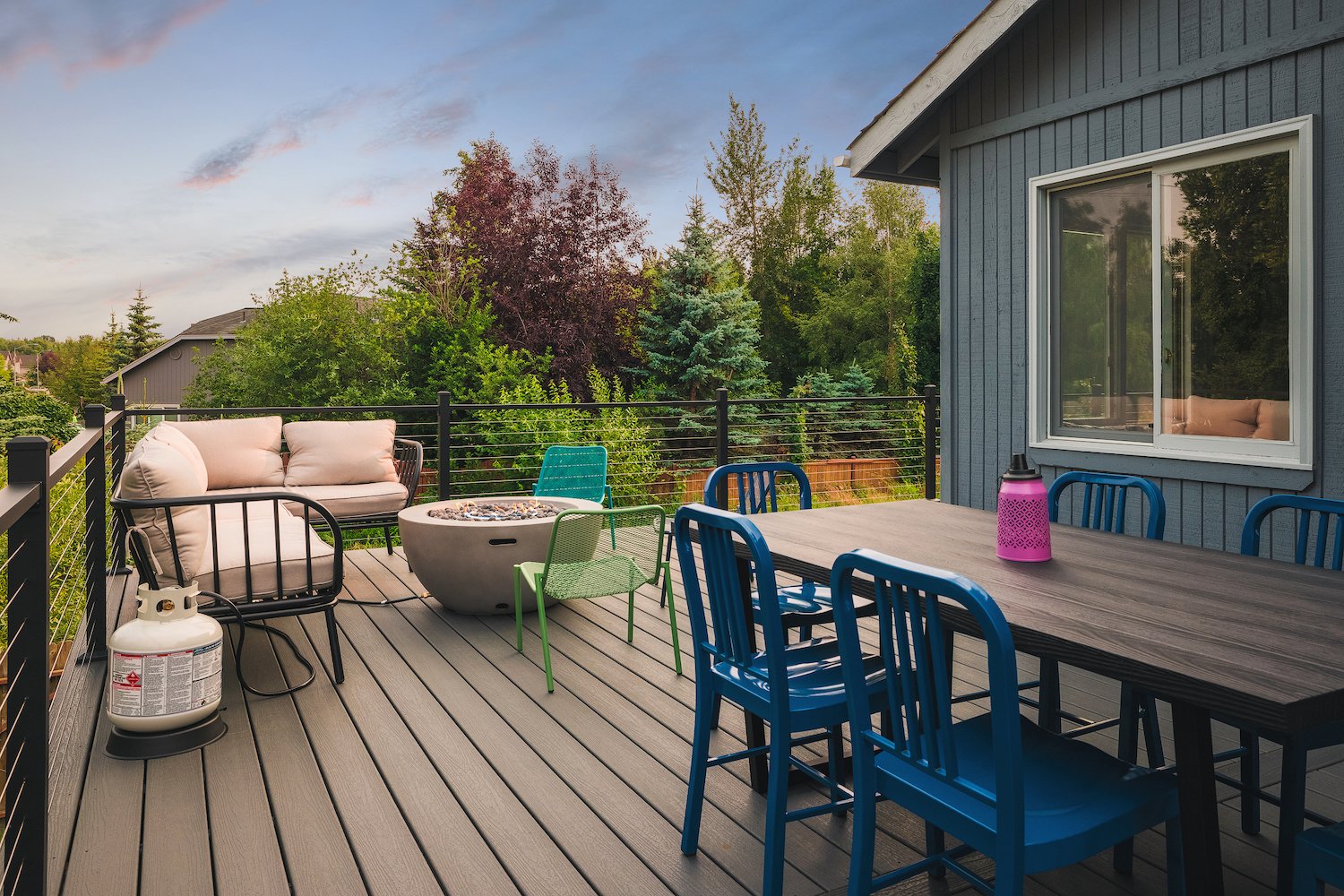 Outdoor trex deck with patio furniture, including a blue dining set, a pink lantern, and a beige sofa with cushions, surrounded by green trees under a cloudy sky.