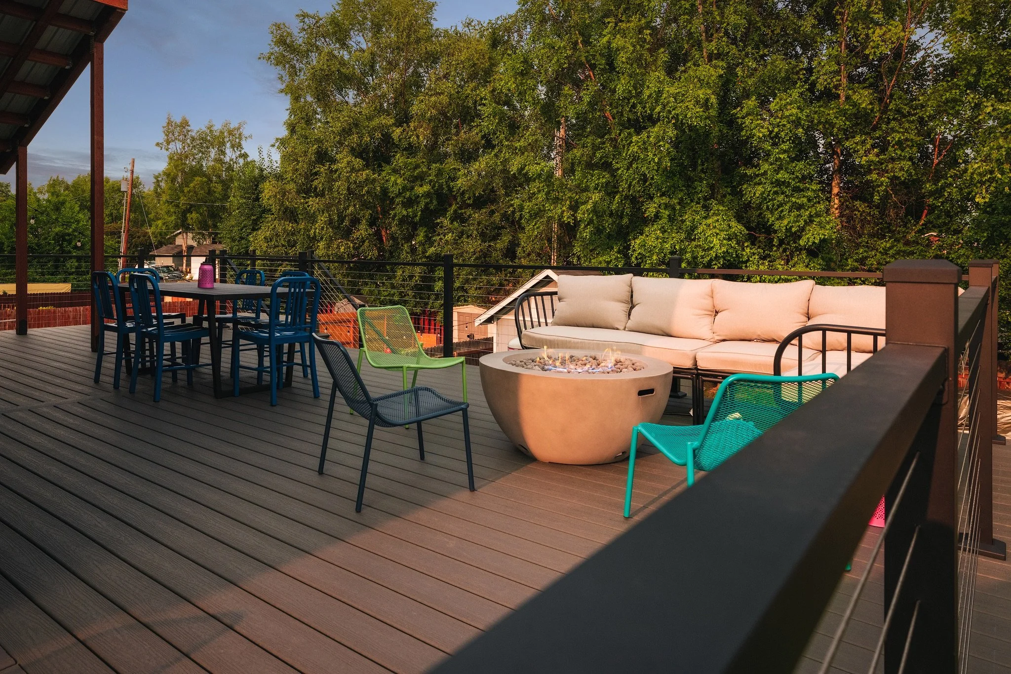 A spacious outdoor balcony with composite decking, featuring a seating area with a long cushioned sofa, colorful metal chairs, a round fire pit table with flames, and a dining table with chairs, surrounded by trees and blue sky.