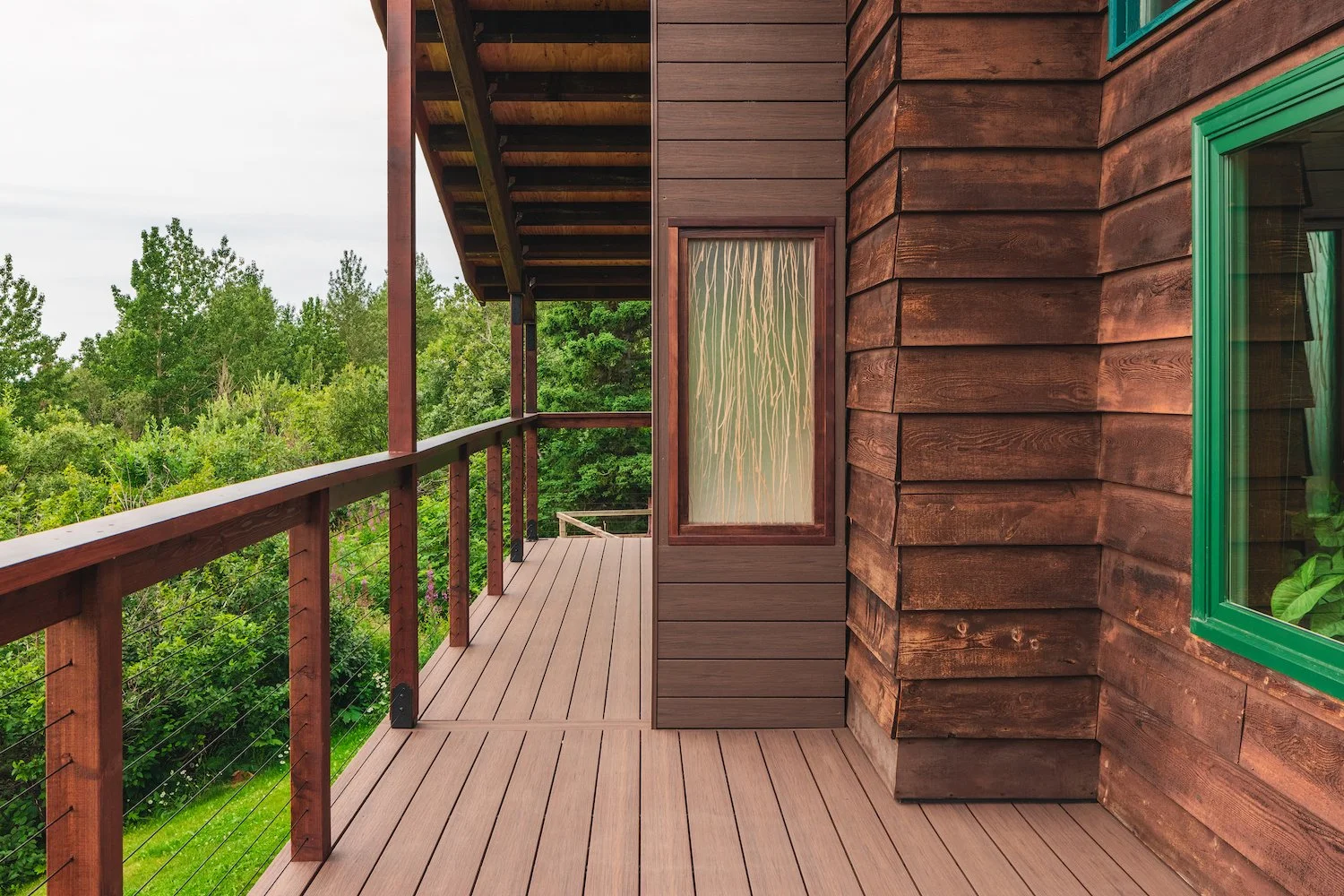 Composite decking balcony with railing, overlooking green trees, attached to a house with brown wooden siding and windows with green frames.