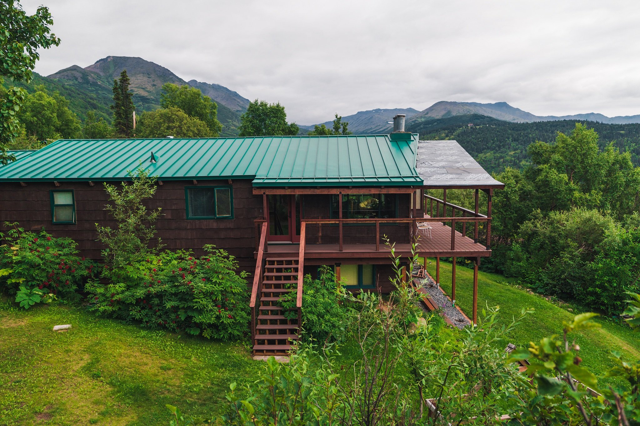 A house with a green metal roof, surrounded by green trees and bushes, with mountains in the background.