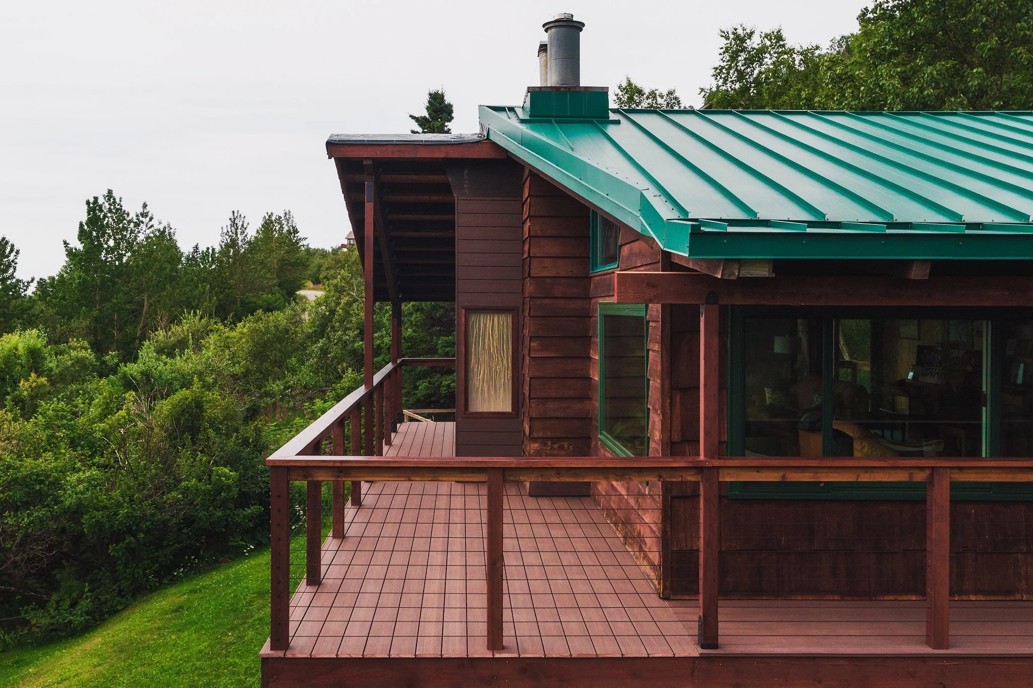 A wooden house with a green metal roof and a balcony surrounded by lush green trees.
