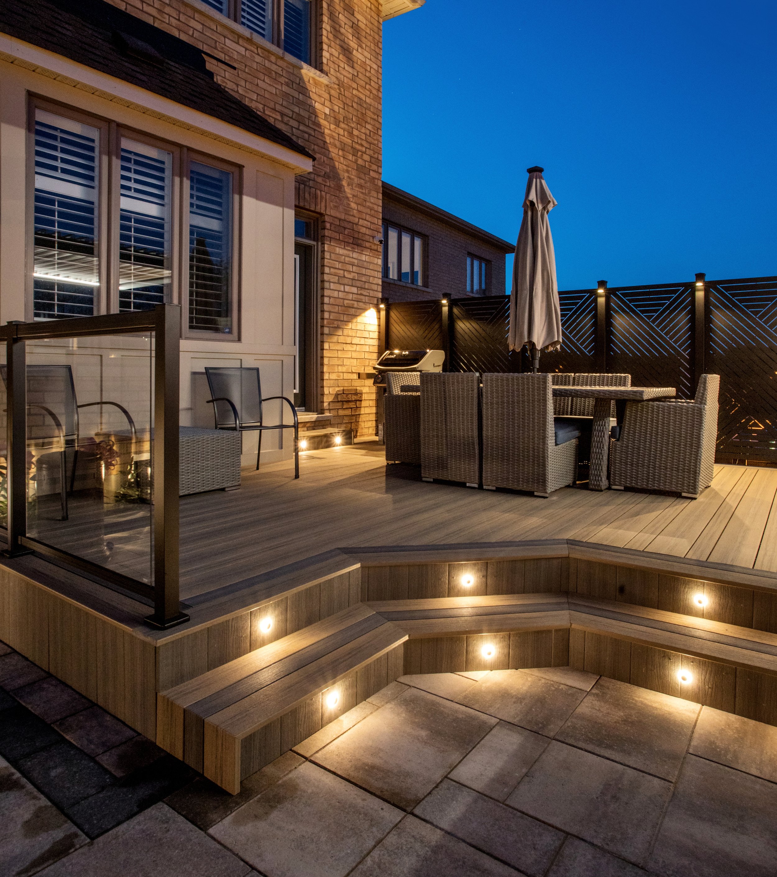 A backyard patio with outdoor furniture, including a dining table with chairs, a grill, and a seating area, illuminated by embedded and wall lighting, with a black privacy fence and brick house exterior visible in the evening.
