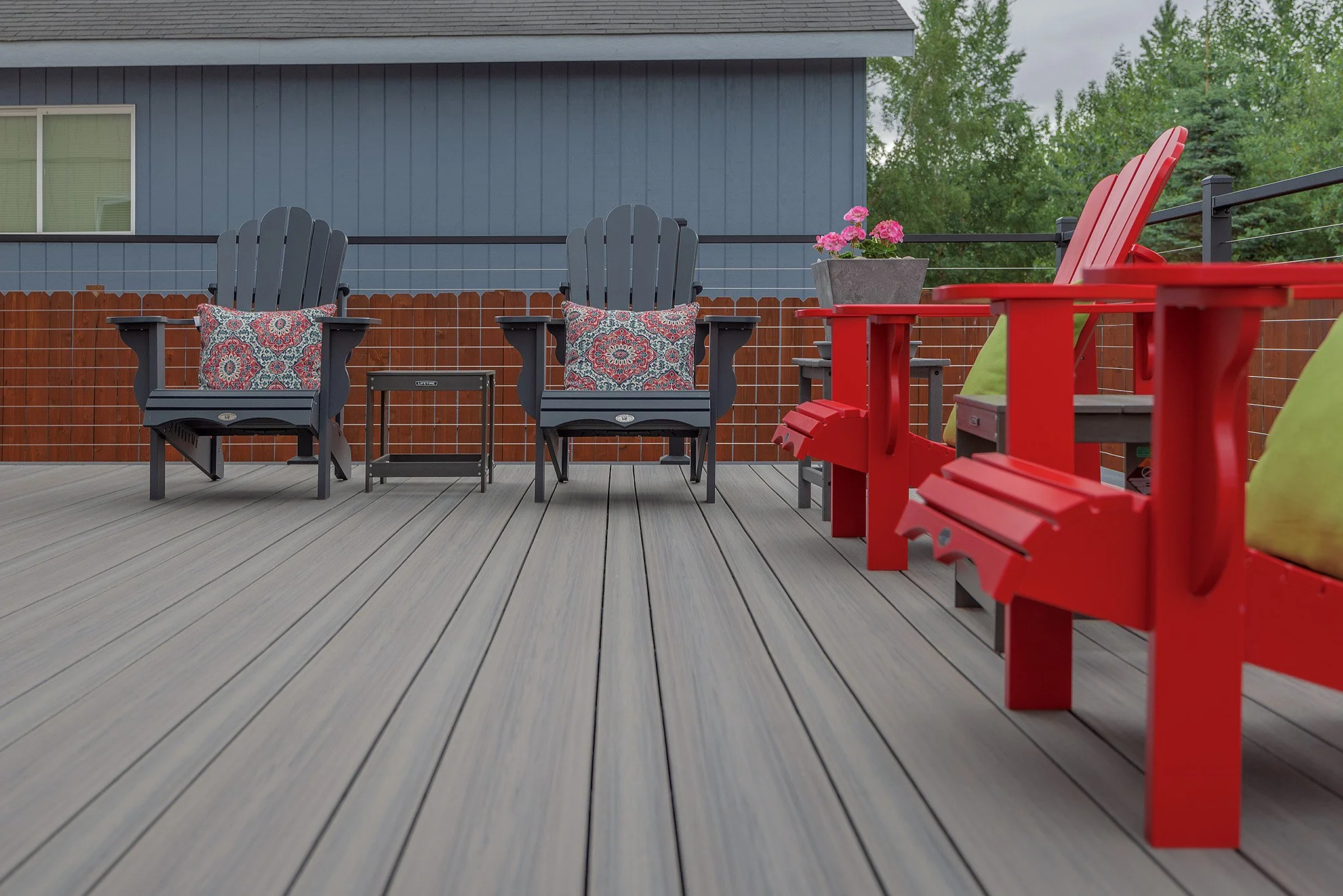 A composite deck with two black Adirondack chairs with patterned pillows, red Adirondack chairs with green pillows, and a small black side table, against a wooden railing and gray house with trees in the background.