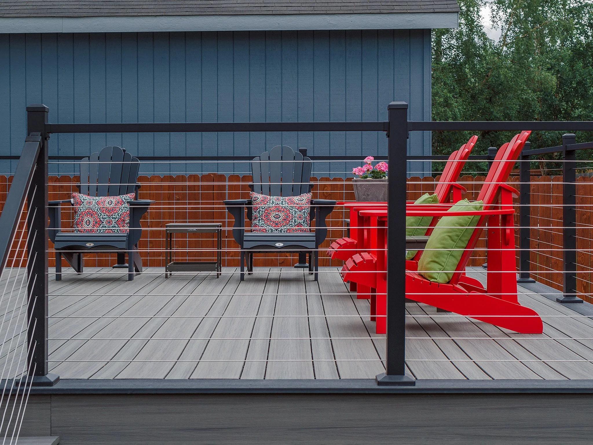 An outdoor deck with black and red Adirondack chairs, small tables, a potted plant with pink flowers, and decorative cushions, enclosed by a black railing.
