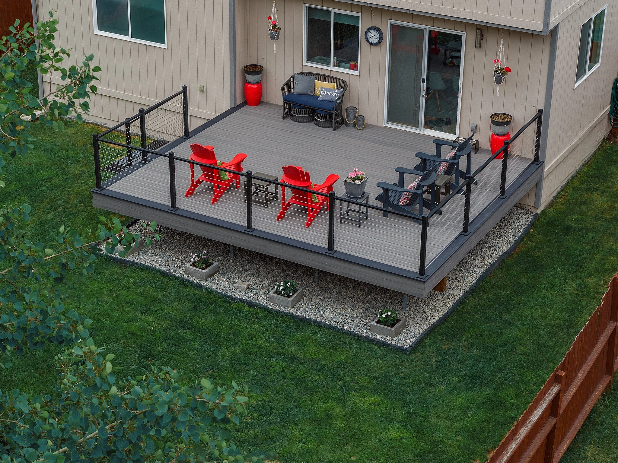 A composite deck attached to a house with outdoor furniture including red and black chairs, a small table with flowers, and a bench. The house has beige siding, a sliding glass door, and windows.