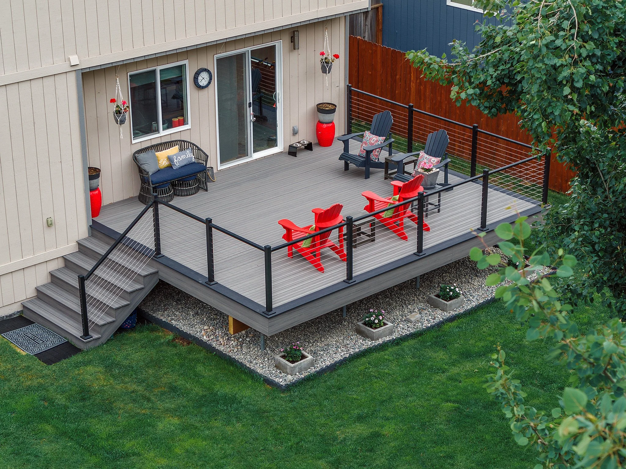 A backyard deck with outdoor furniture, including two red Adirondack chairs and two black Adirondack chairs with floral cushions, enclosed by a black metal railing, with a grassy yard and trees in the background.