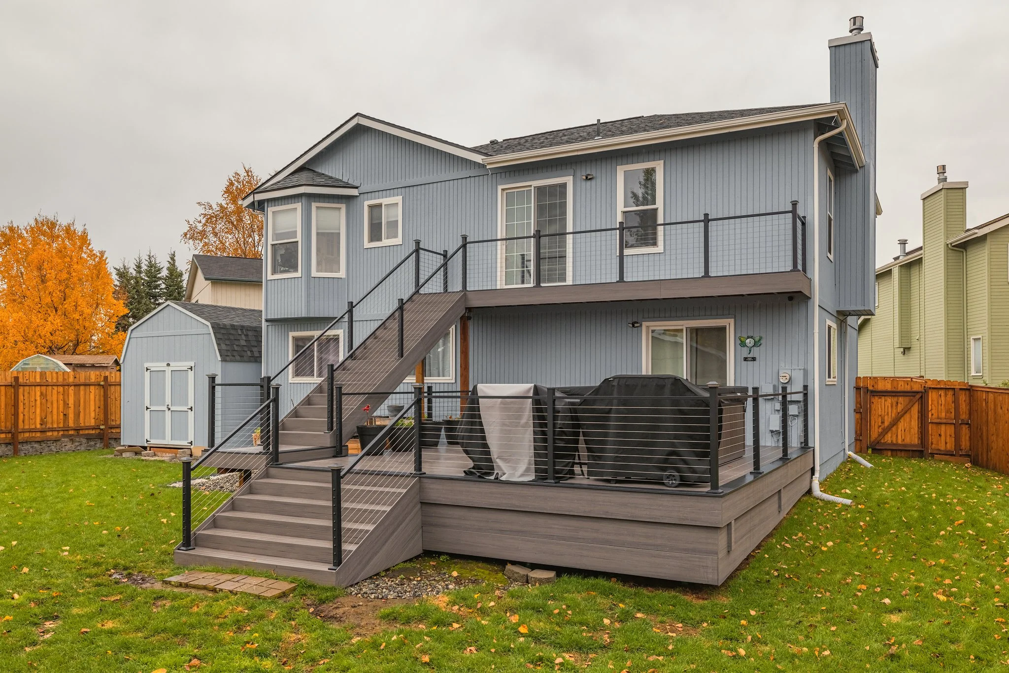 Backyard view of a two-story blue house with a deck, outdoor grill covered with a black cover, and stairs leading down to a grassy yard, during overcast weather with trees showing fall colors in the background.