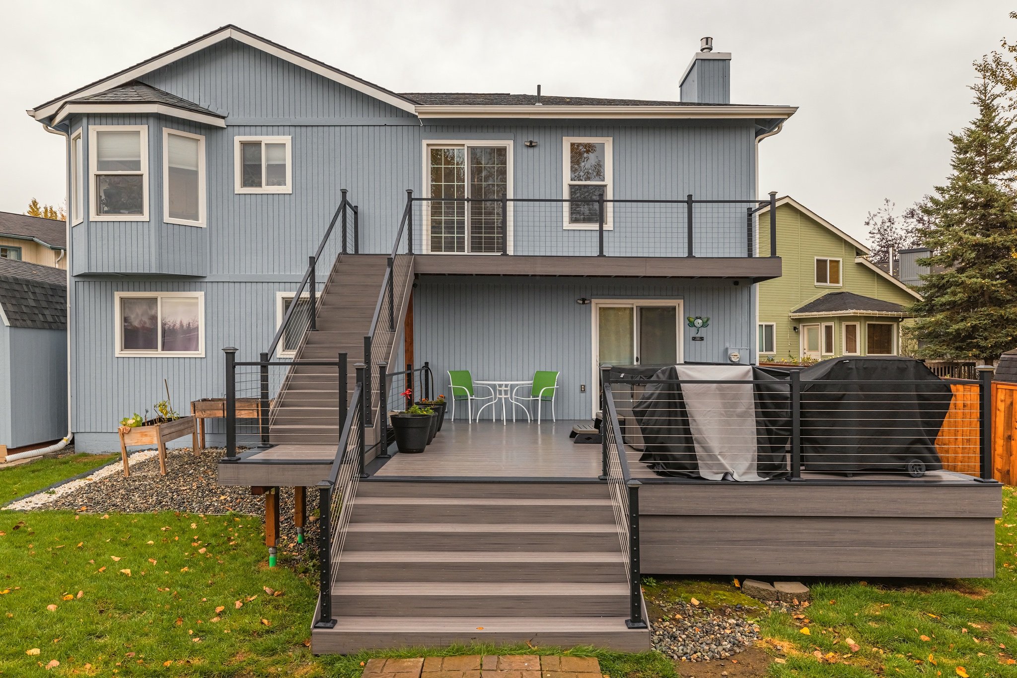 Backyard view of a two-story blue house with composite decking deck, stairs, and patio furniture, including a round table and two chairs, along with covered grill items, in a suburban neighborhood with neighboring houses and trees.