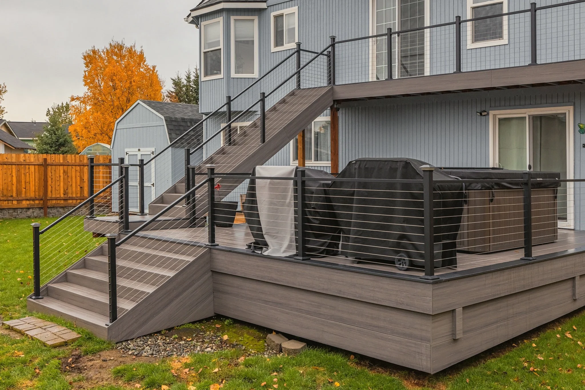 Backyard patio with stairs, black railing, outdoor furniture, and covered grill, attached to a blue house with large windows and a wooden fence in the background.