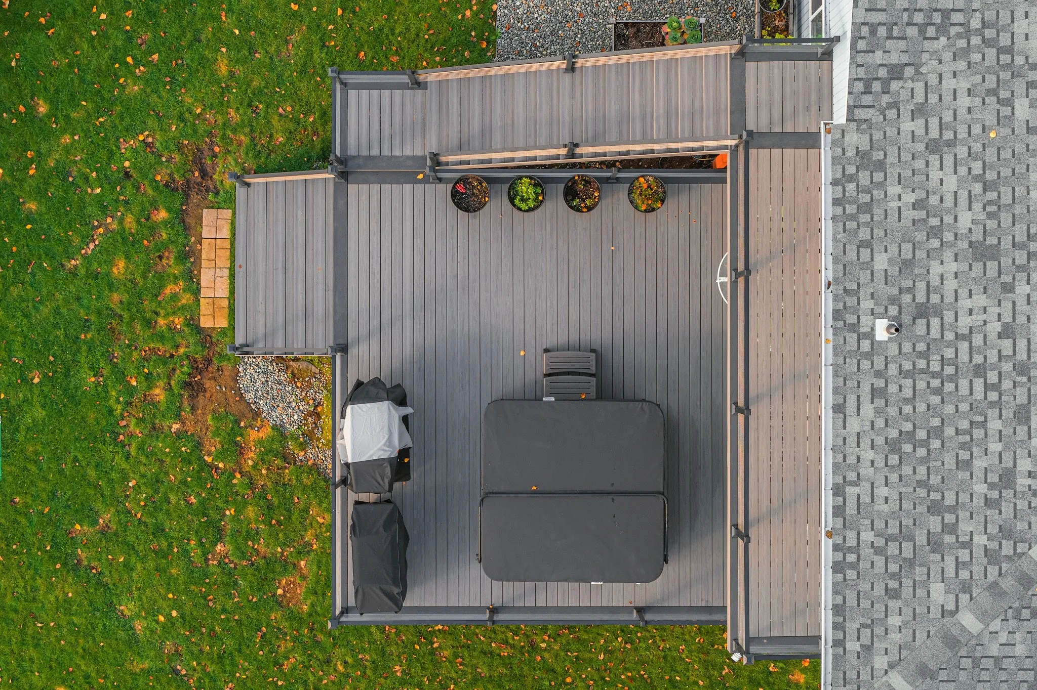 An aerial view of a backyard deck with outdoor furniture, potted plants, a barbecue grill, and a volleyball net, surrounded by a grassy yard with fallen leaves and a patio area with gray paving stones.