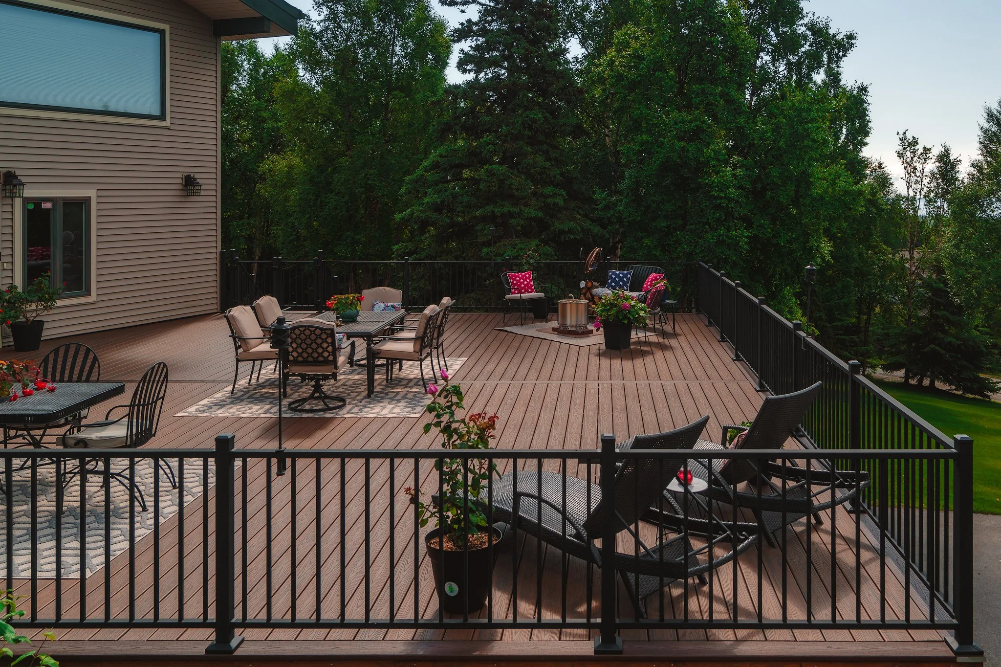A spacious outdoor wooden deck attached to a house, furnished with multiple chairs, tables, potted plants, and decorated with cushions and flowers, surrounded by lush green trees.