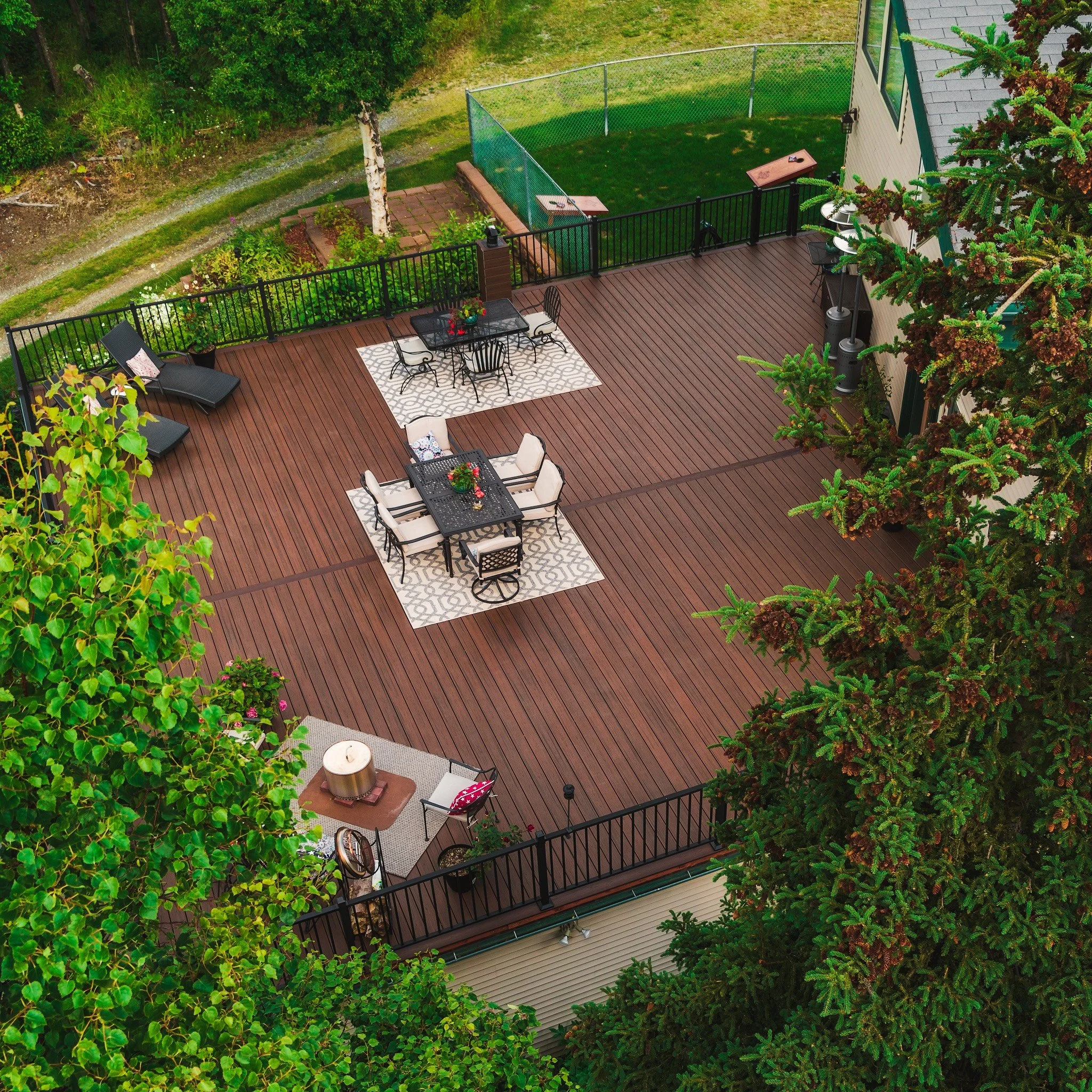 A spacious outdoor wooden deck with black iron furniture, including tables, chairs, and lounge chairs, surrounded by trees and greenery, with a fenced tennis court in the background.