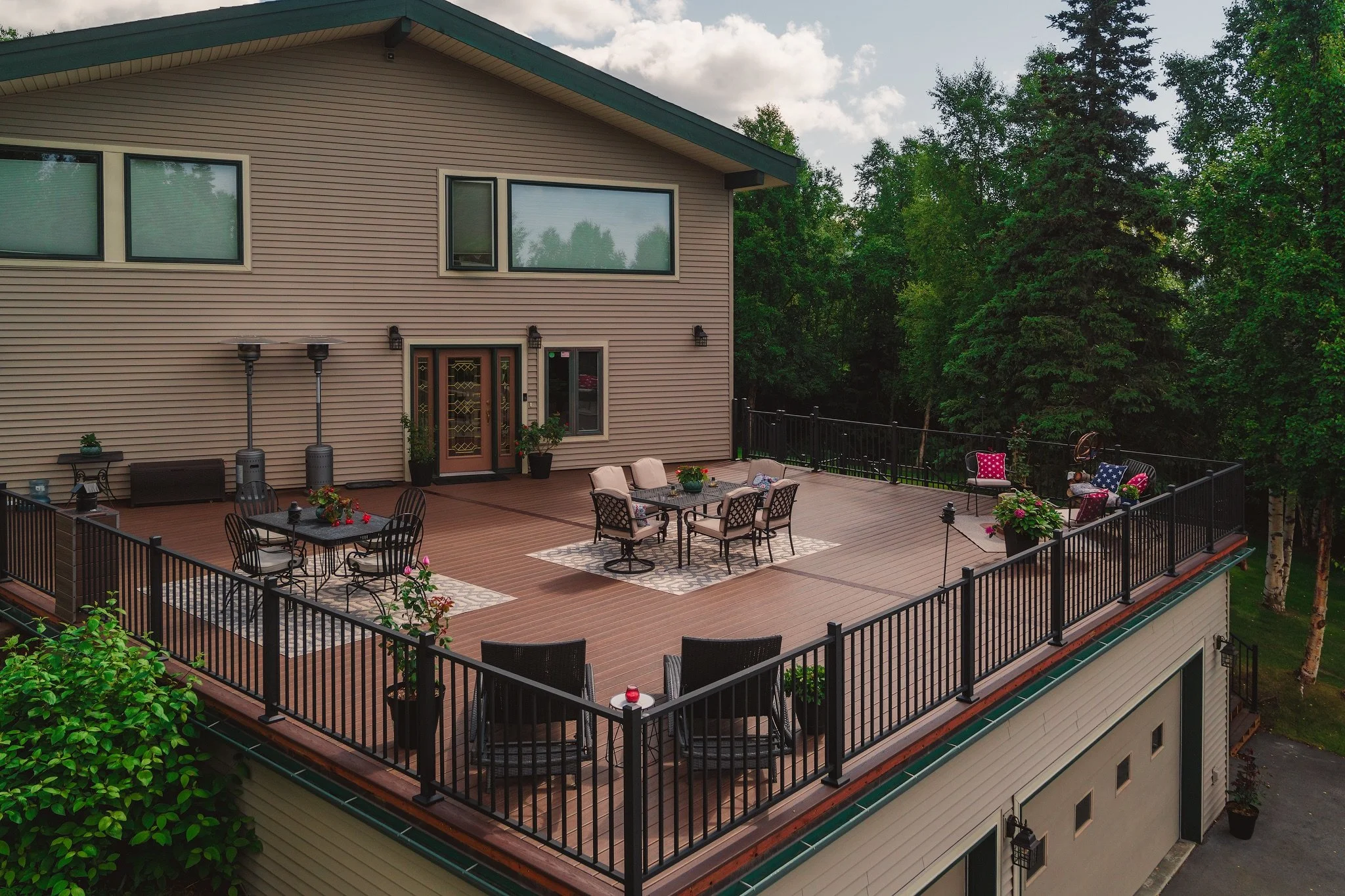 A spacious outdoor rooftop deck with patio furniture, potted plants, and string lights, surrounded by a black metal railing and greenery in the background.