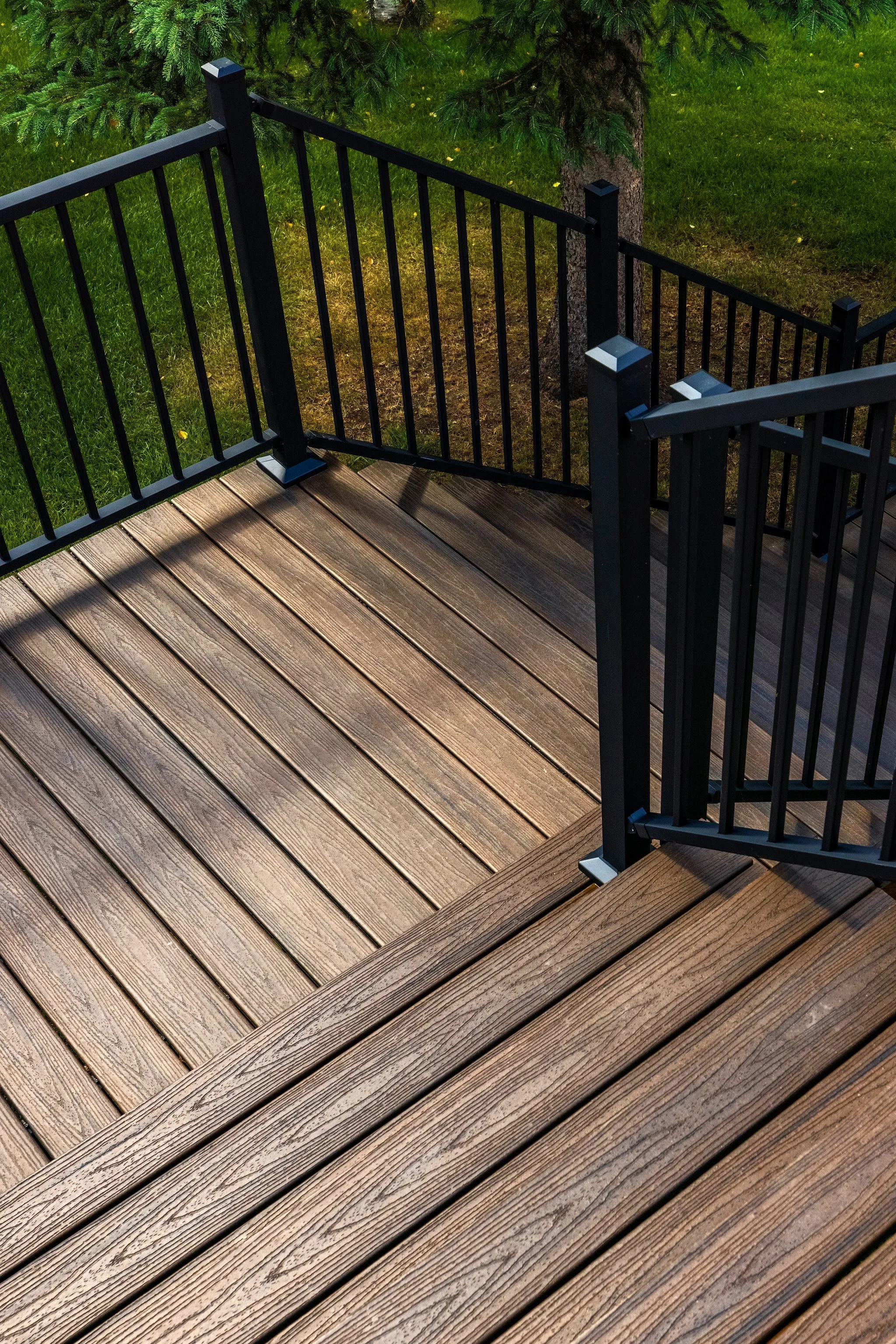 A wooden deck with black metal railing around the edges, adjacent to a grassy yard with trees.