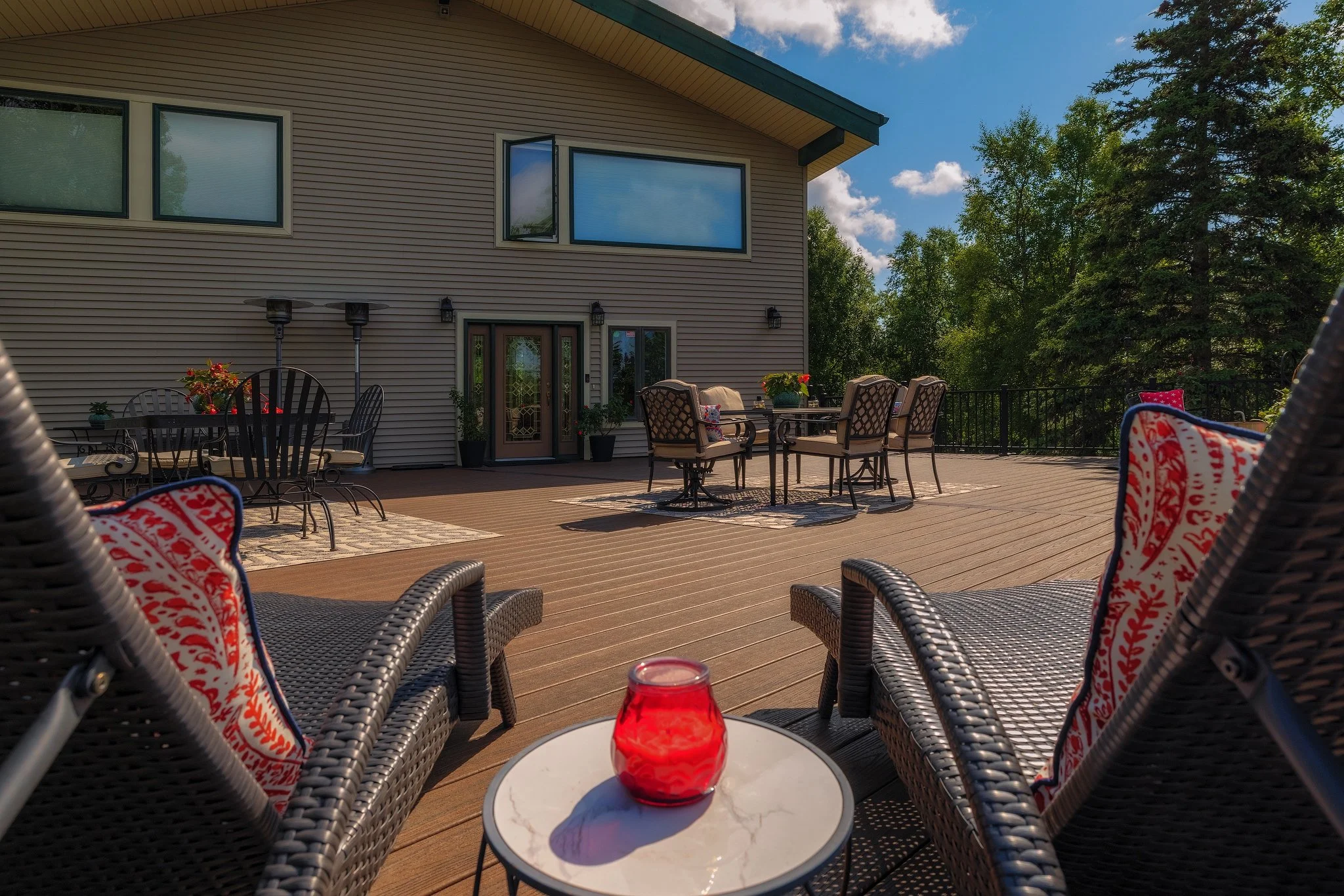 Spacious outdoor deck with wicker chairs, colorful cushions, and a coffee table with a red candle, attached to a house with large windows and surrounded by trees on a sunny day.