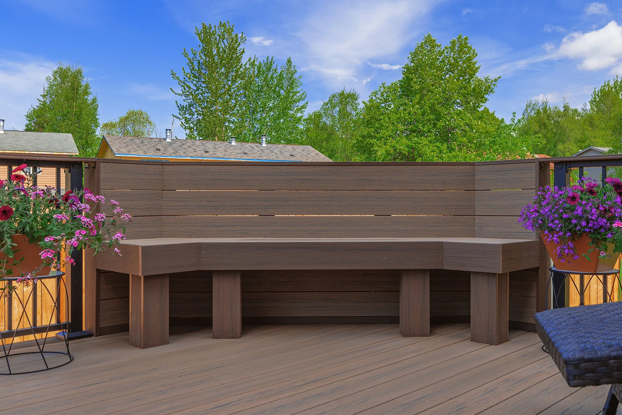 A backyard deck with a wooden bench, potted pink and purple flowers, and a privacy fence made of horizontal wooden planks. Green trees and a blue sky with some clouds are in the background.