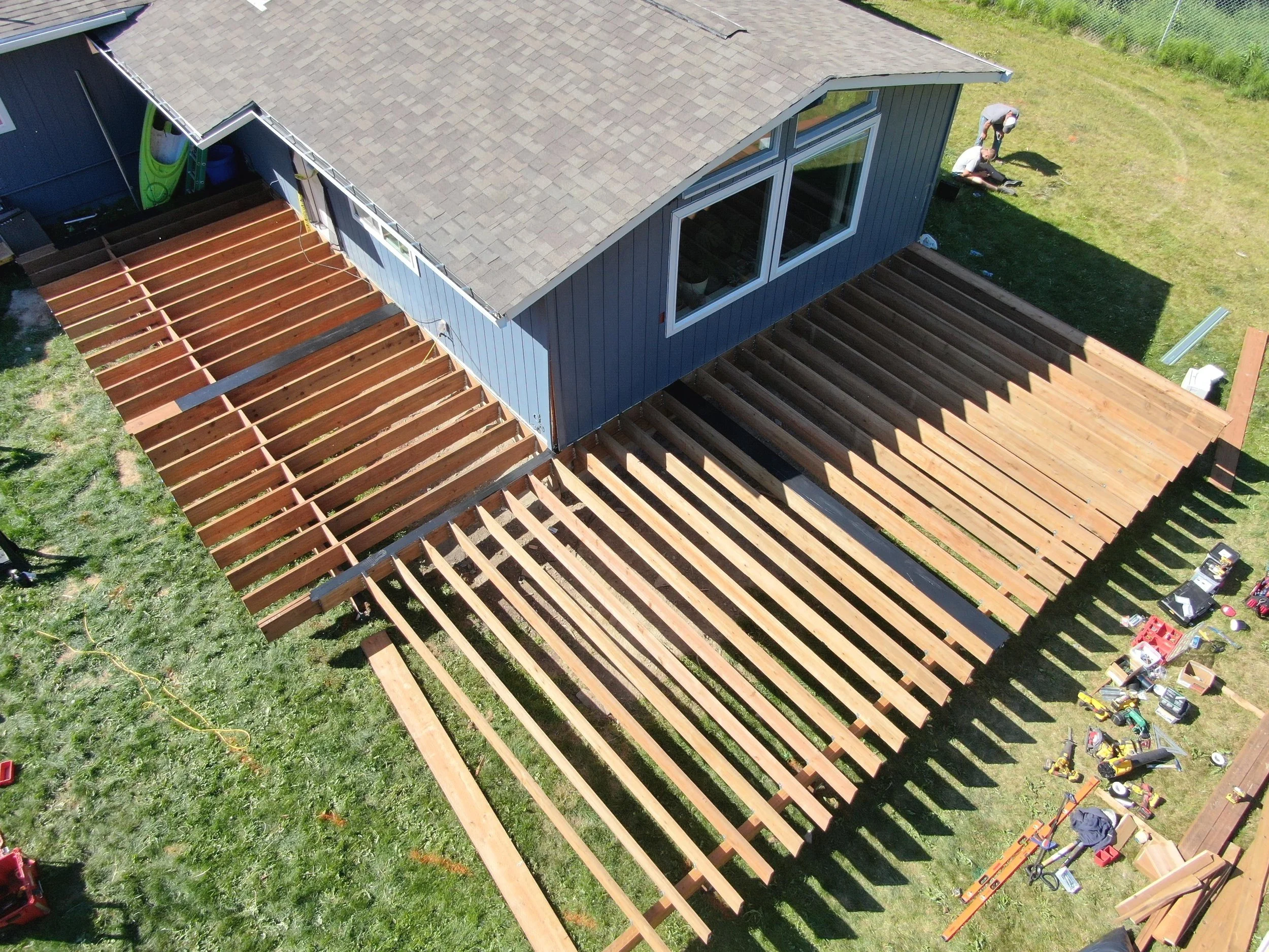 Aerial view of a house under construction with wooden deck framing on the back and side, construction tools and materials scattered on the lawn.
