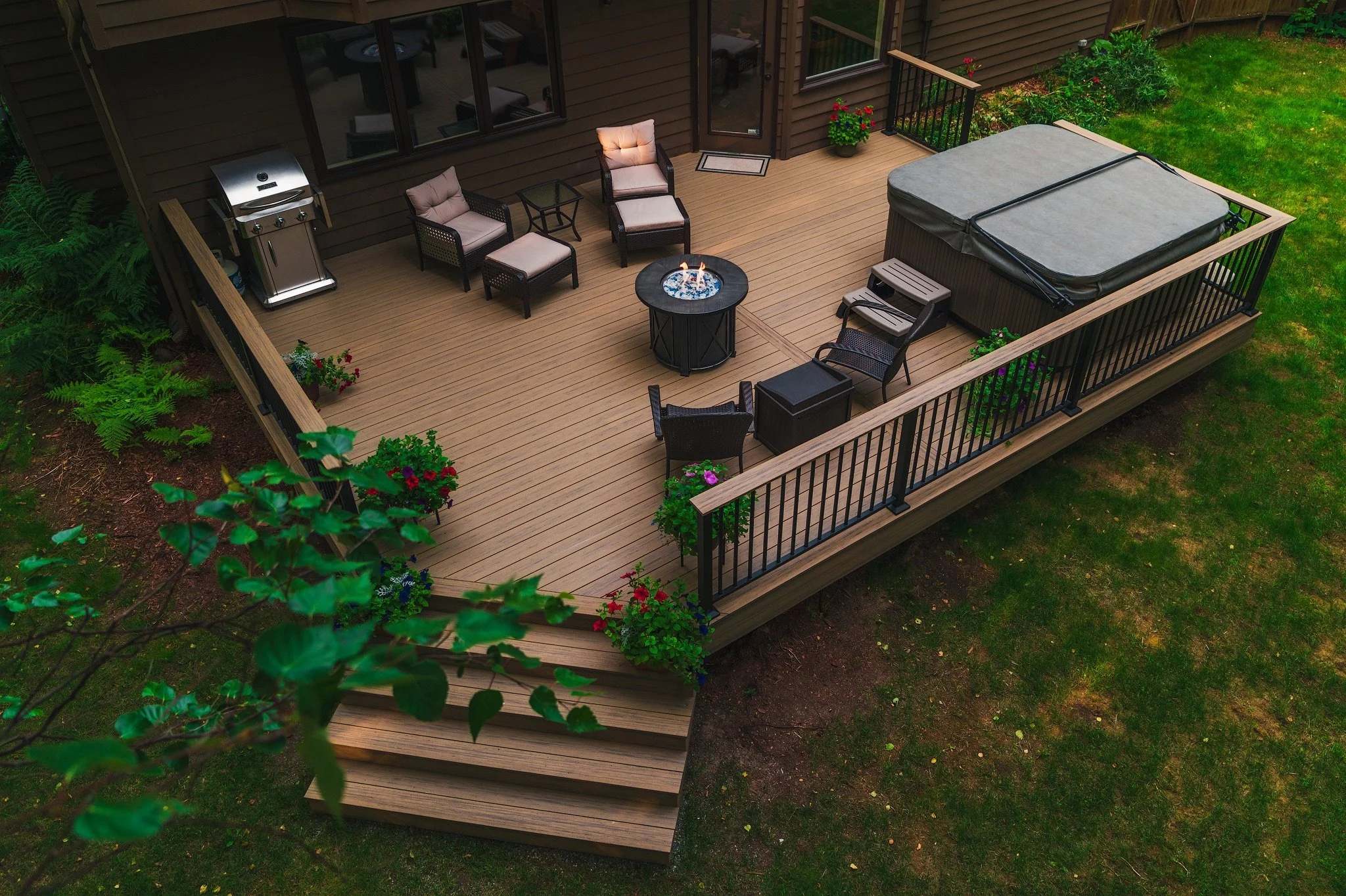 A wooden deck attached to a house with outdoor furniture, a hot tub, and a fire pit surrounded by a black metal railing. The deck has potted plants and is accessed by stairs from a grassy yard.