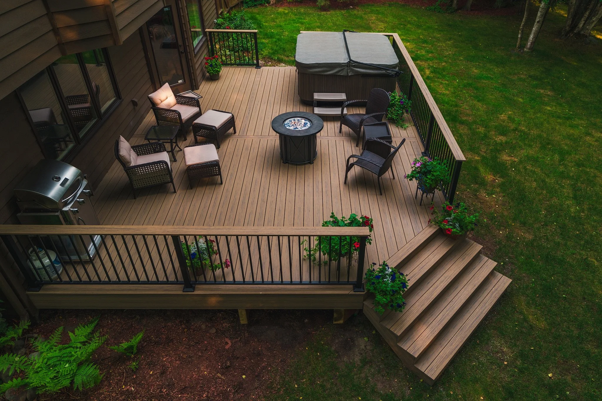An outdoor wooden deck with furniture including a gas grill, several chairs, and potted flowers, surrounded by green grass and trees.