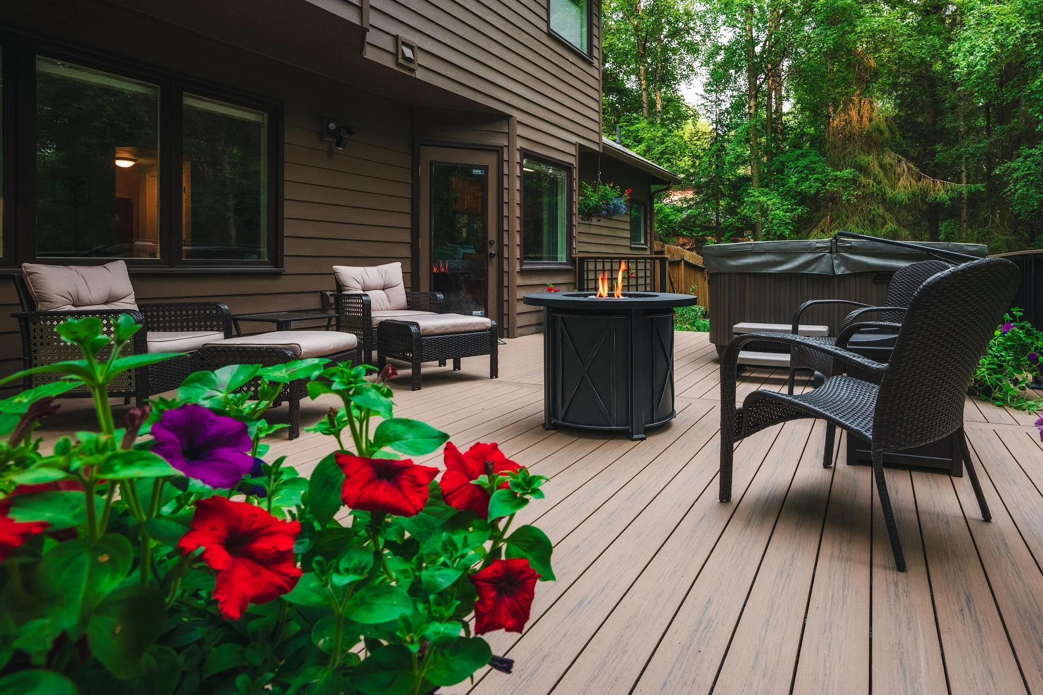 Back deck of a house with outdoor seating, a fire pit, potted flowers, and surrounded by green trees.