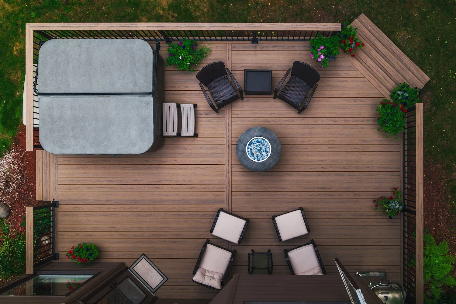 Top-down view of a wooden deck patio with two black chairs and two matching white cushions, a small firepit table, a covered hot tub, a wooden bench, and various potted plants with colorful flowers, surrounded by grass and garden soil.