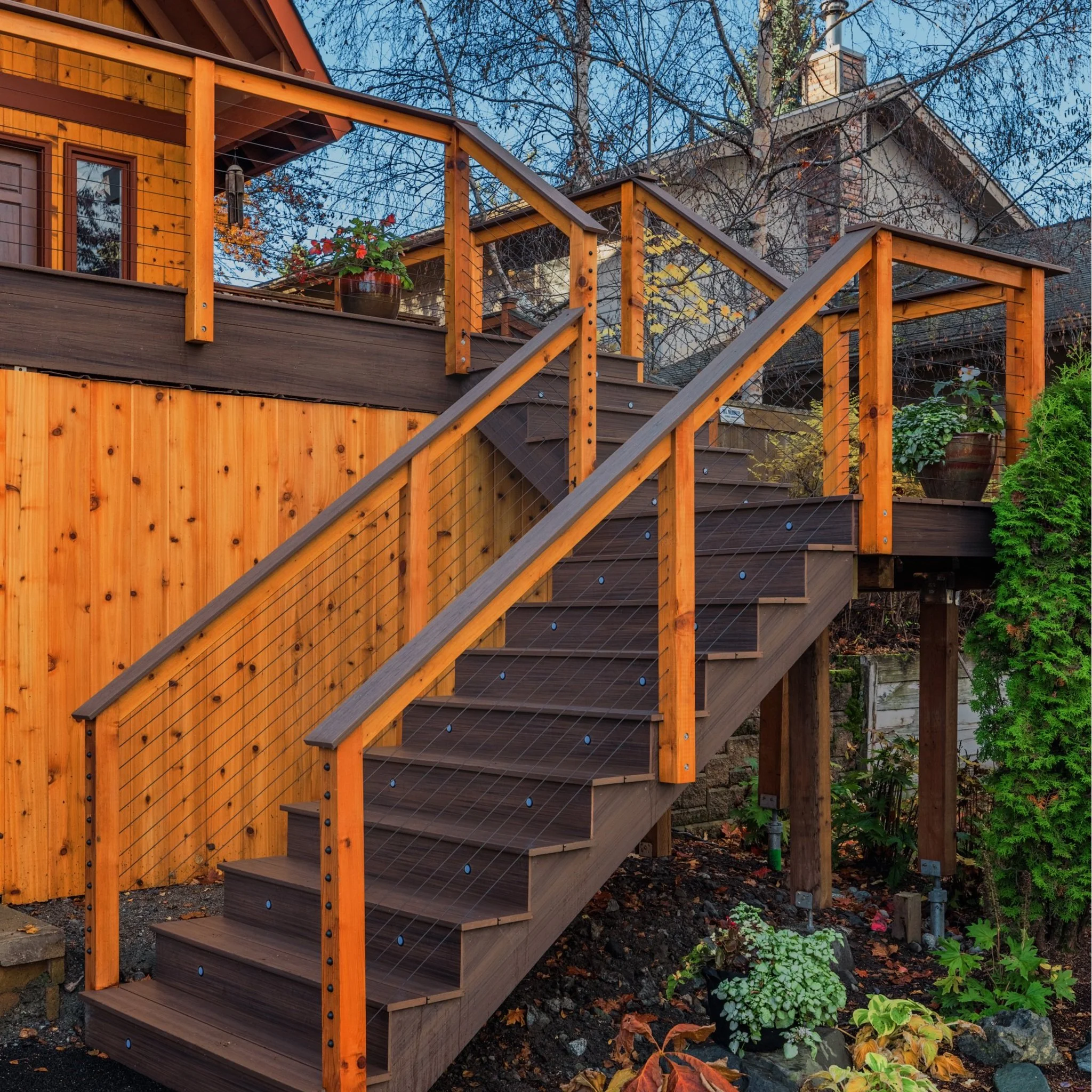 Wooden staircase with safety wires and orange railing leading up to a deck with wooden railing and potted plants, backyard with trees and neighboring house in background.