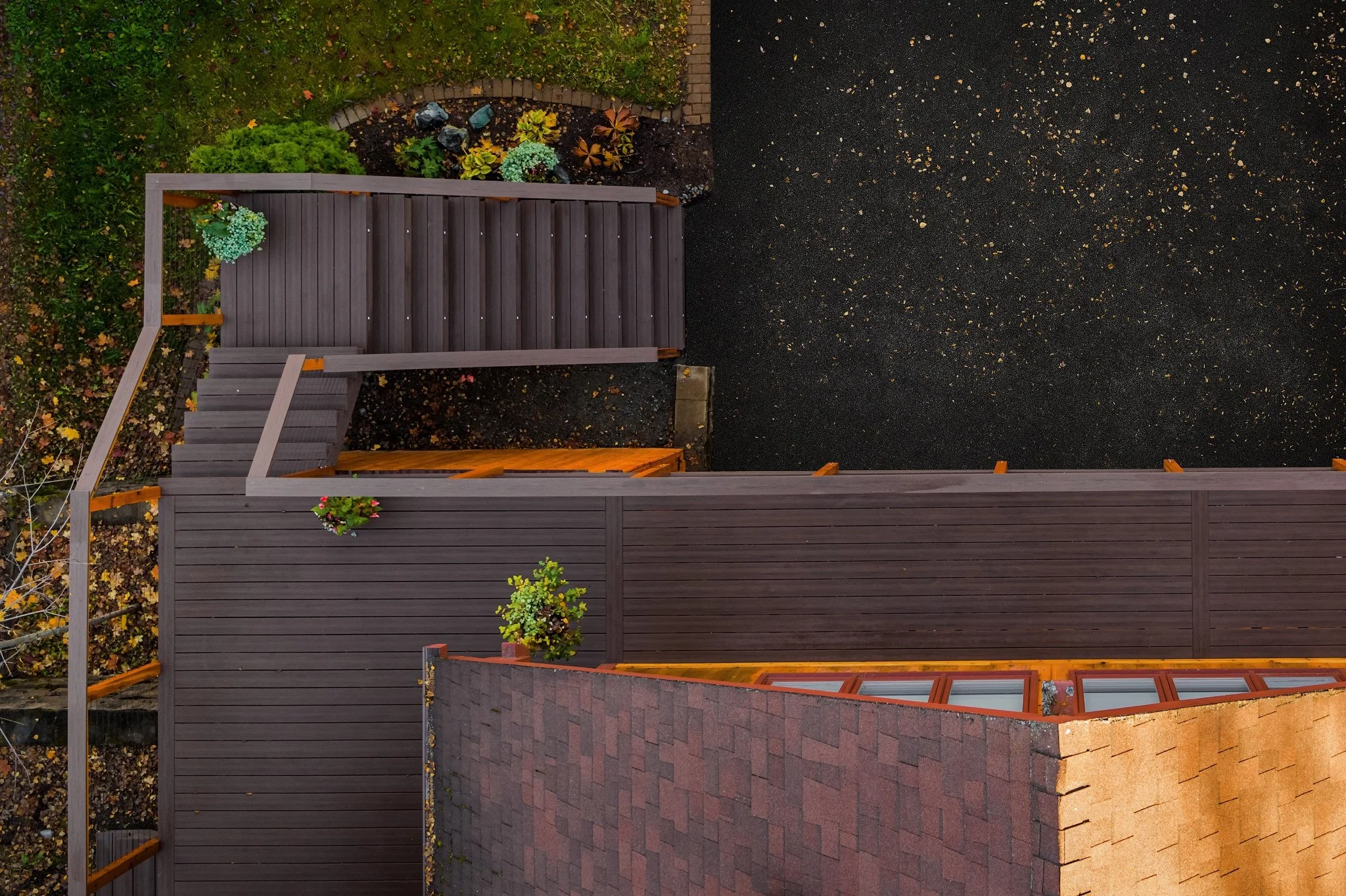 A view from above of a backyard showing a wooden deck, a paved patio, a black asphalt area, a garden with plants, and a brick house roof. There are potted plants and fallen leaves.