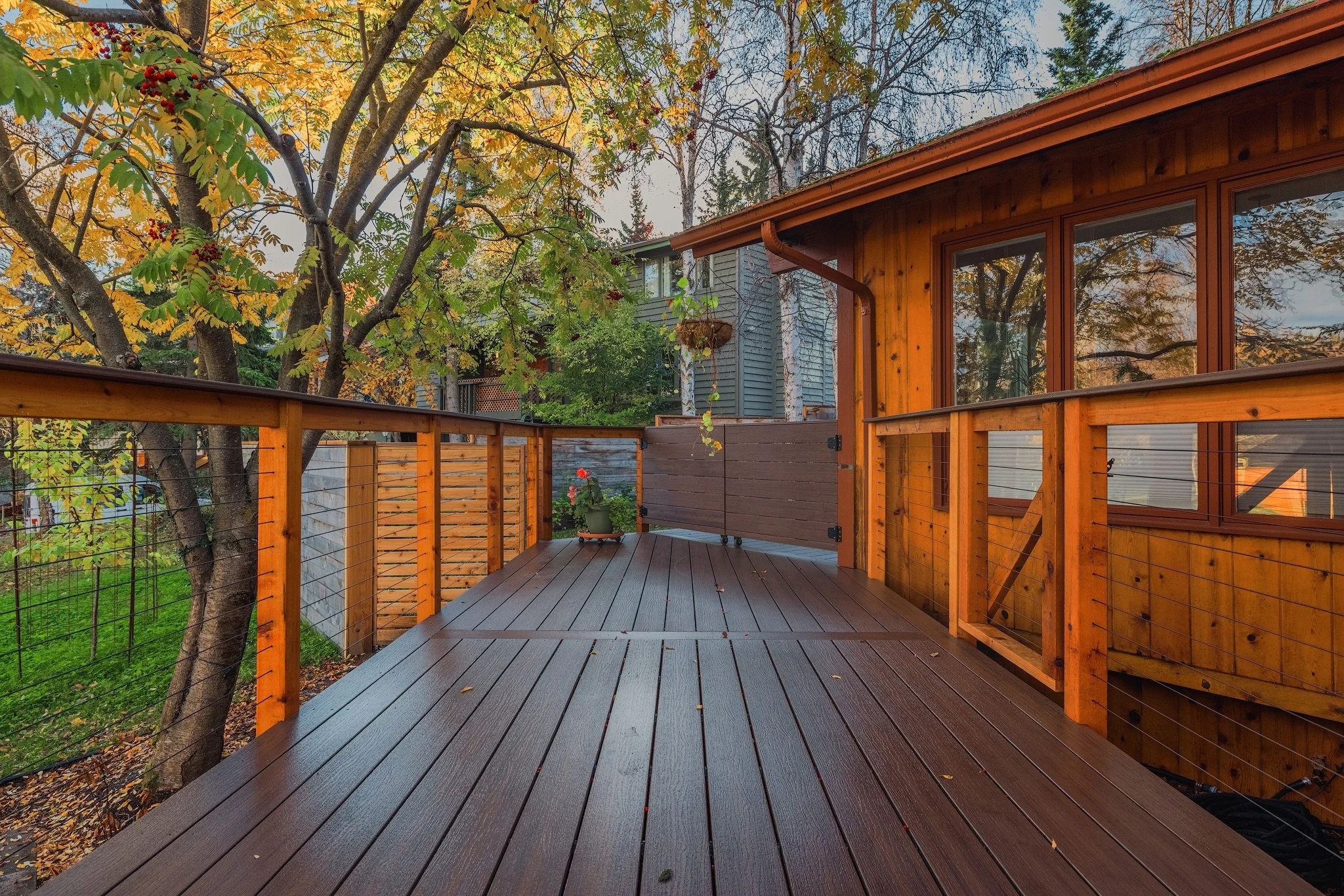Wooden deck with railing attached to a house, surrounded by trees with fall foliage and a garden with a potted plant, in a neighborhood with other houses visible.