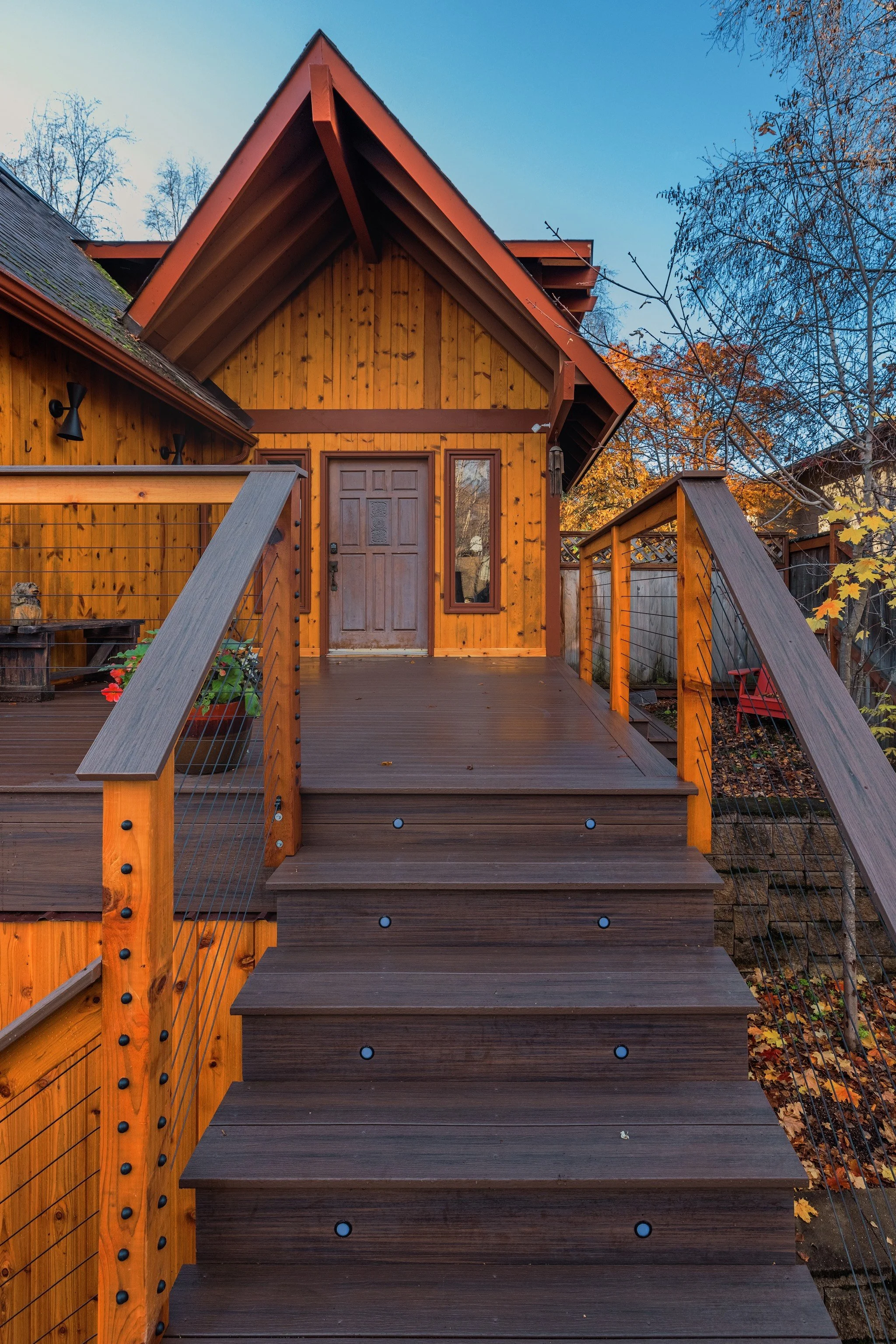 Front view of a wooden house with a staircase leading up to the front door, surrounded by trees with autumn leaves.