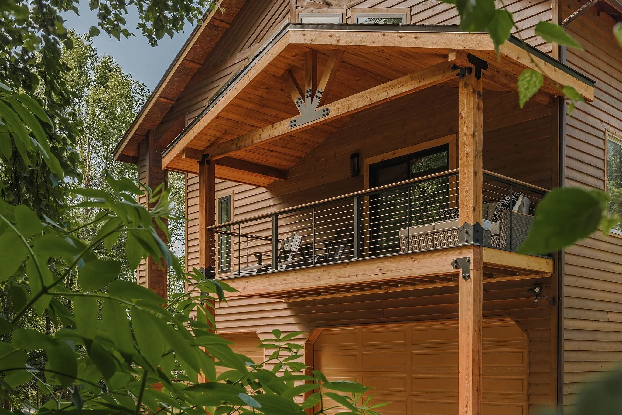 Wooden house with a balcony, black railing, outdoor furniture, surrounded by green trees.