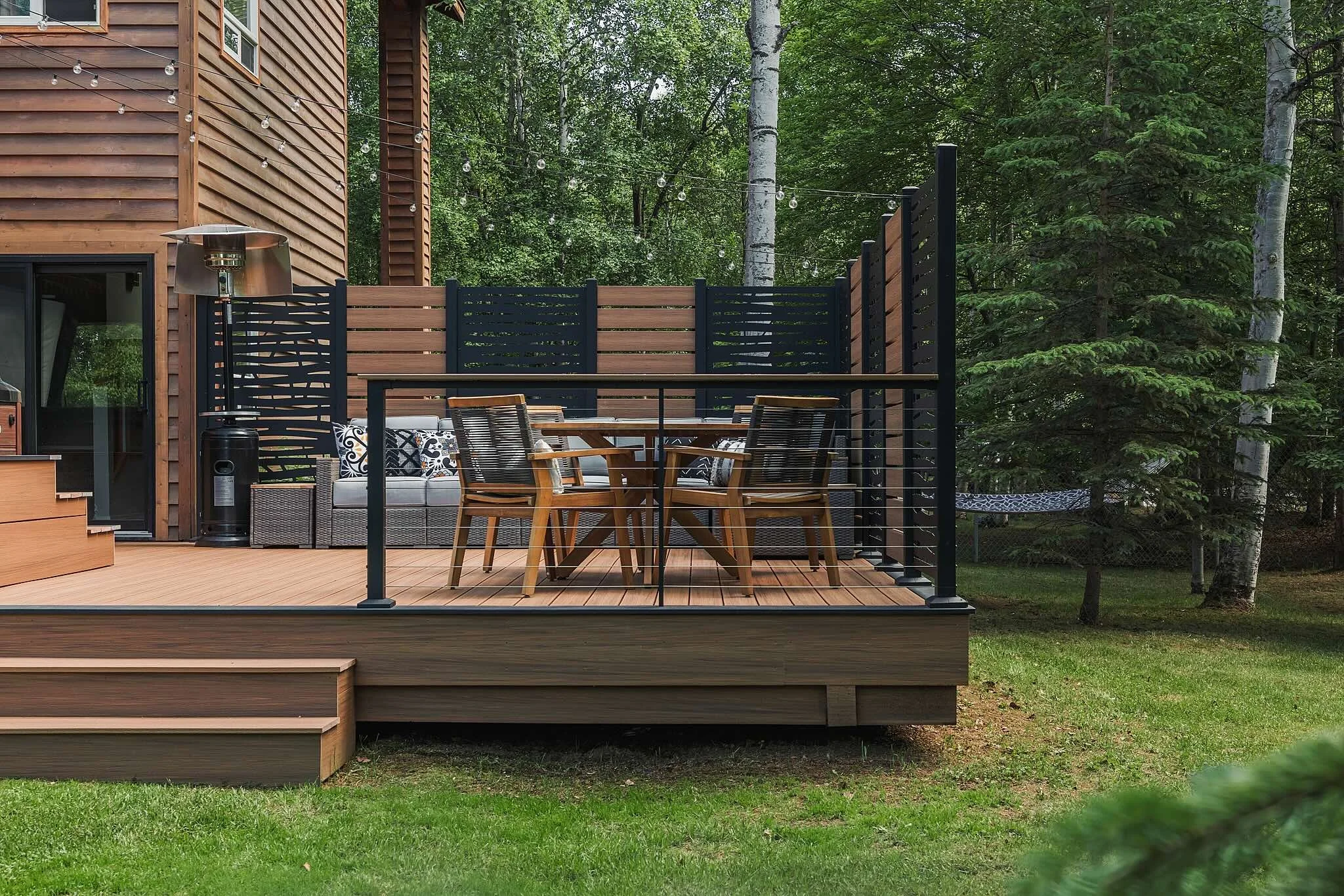 A wooden deck with black railings attached to a house surrounded by trees, featuring outdoor furniture including a table, chairs, a cushioned lounge, and a heater, with a hammock in the background.