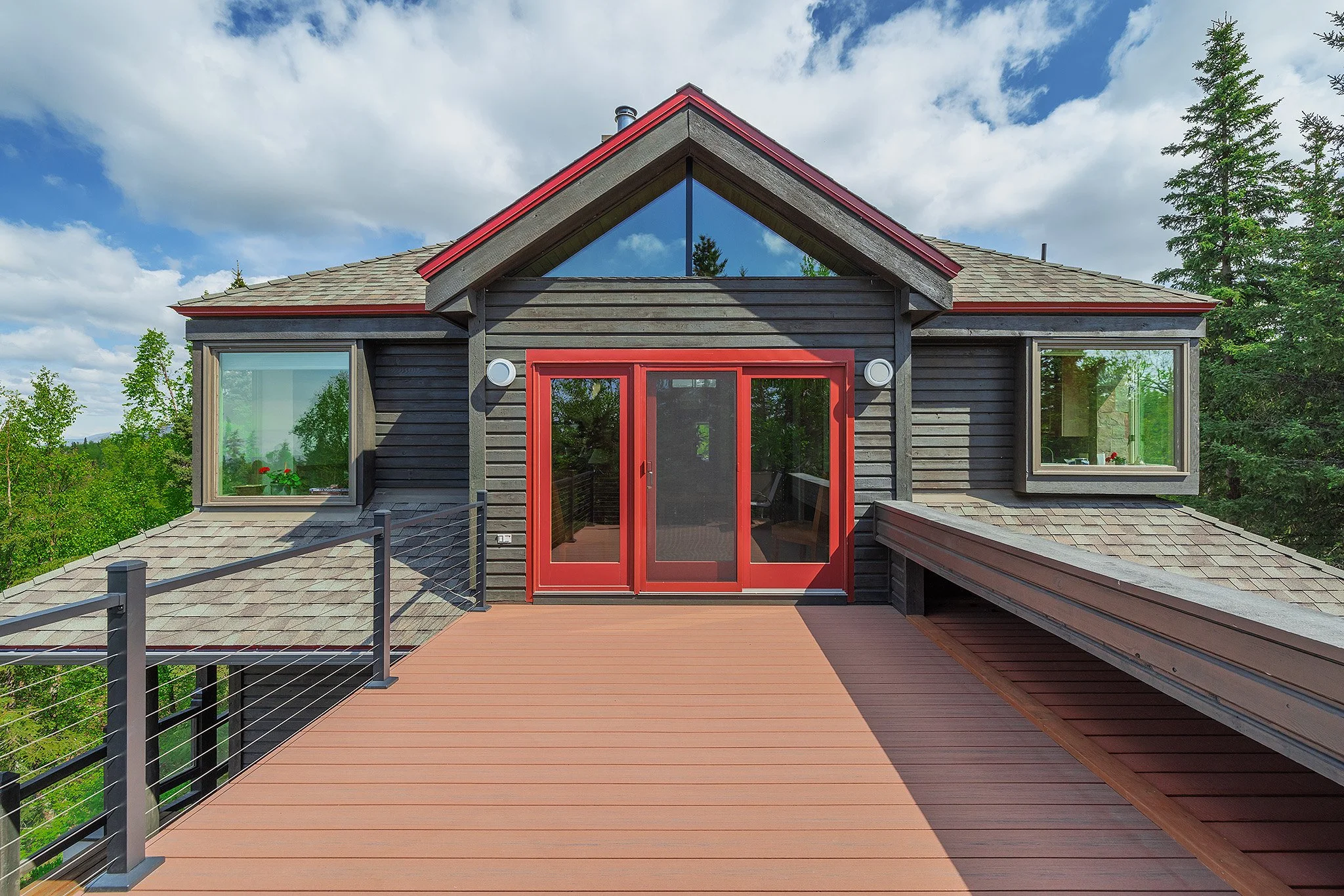 View of a house with a wooden deck, large red double doors, and three windows, surrounded by green trees under a partly cloudy sky.