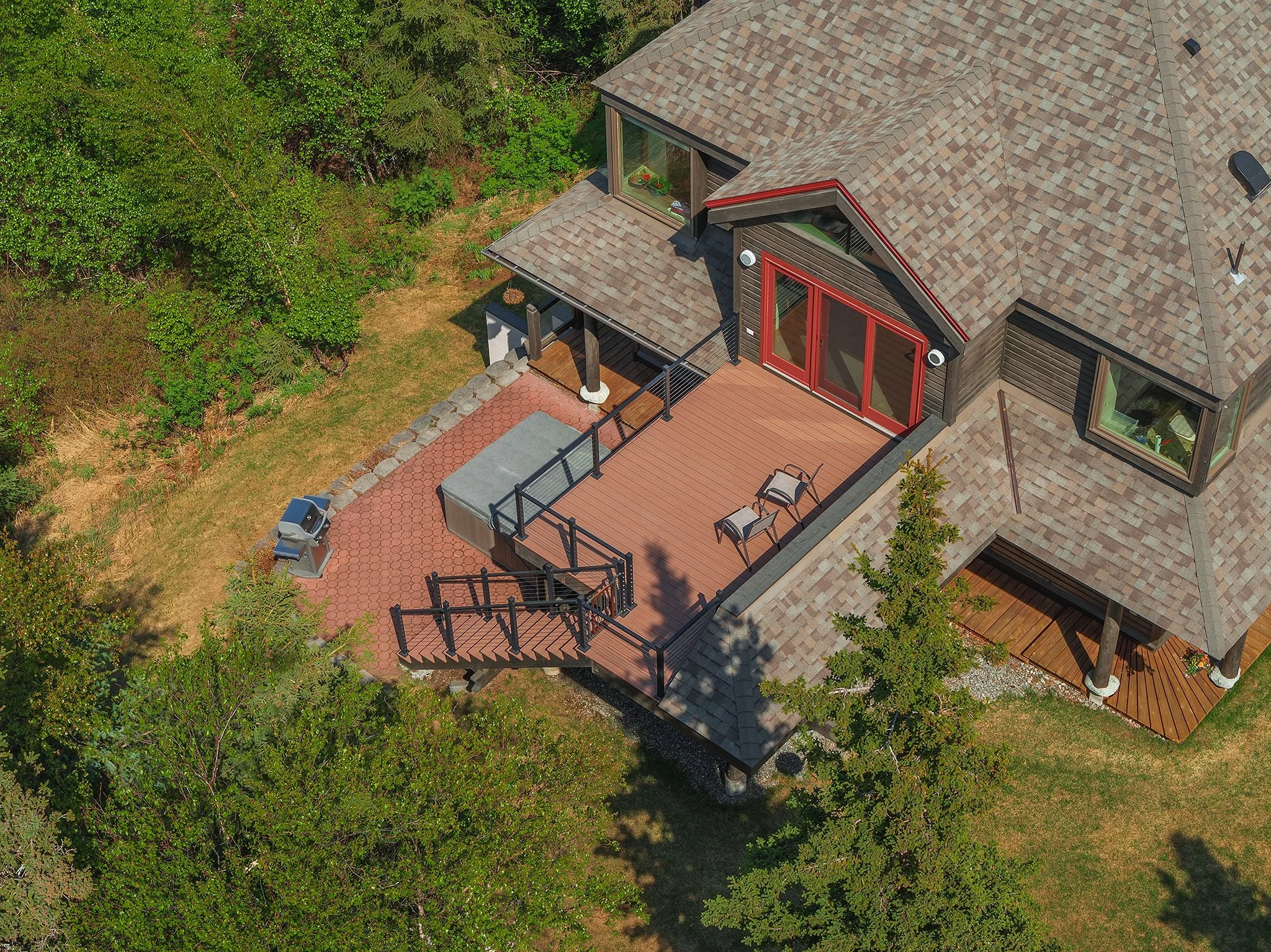Aerial view of a house with a large wooden deck, red sliding doors, and multiple windows. The deck has two chairs, a grill, and is surrounded by trees and greenery.