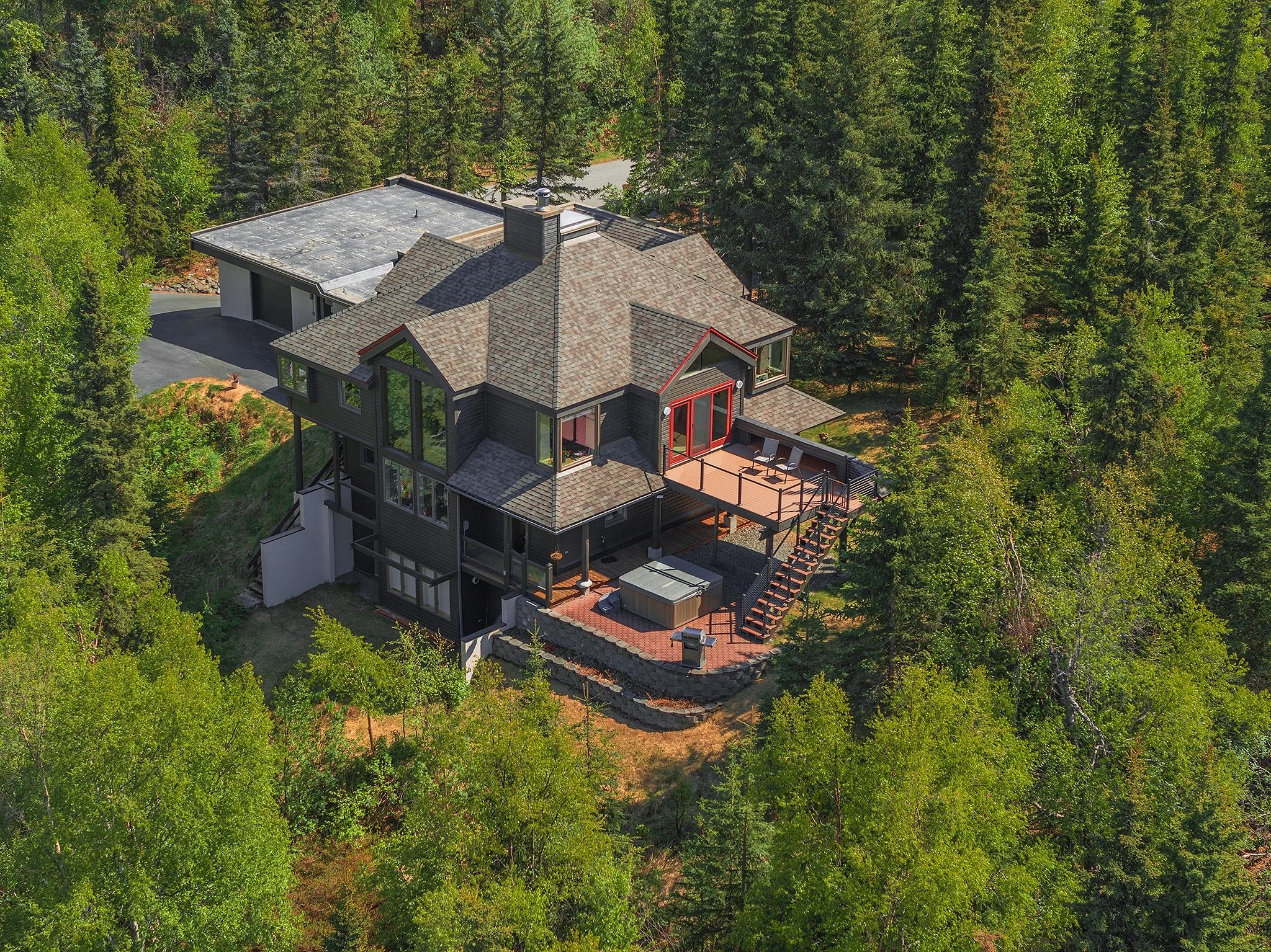 A large house with multiple levels, surrounded by dense green trees, with a deck and outdoor furniture.