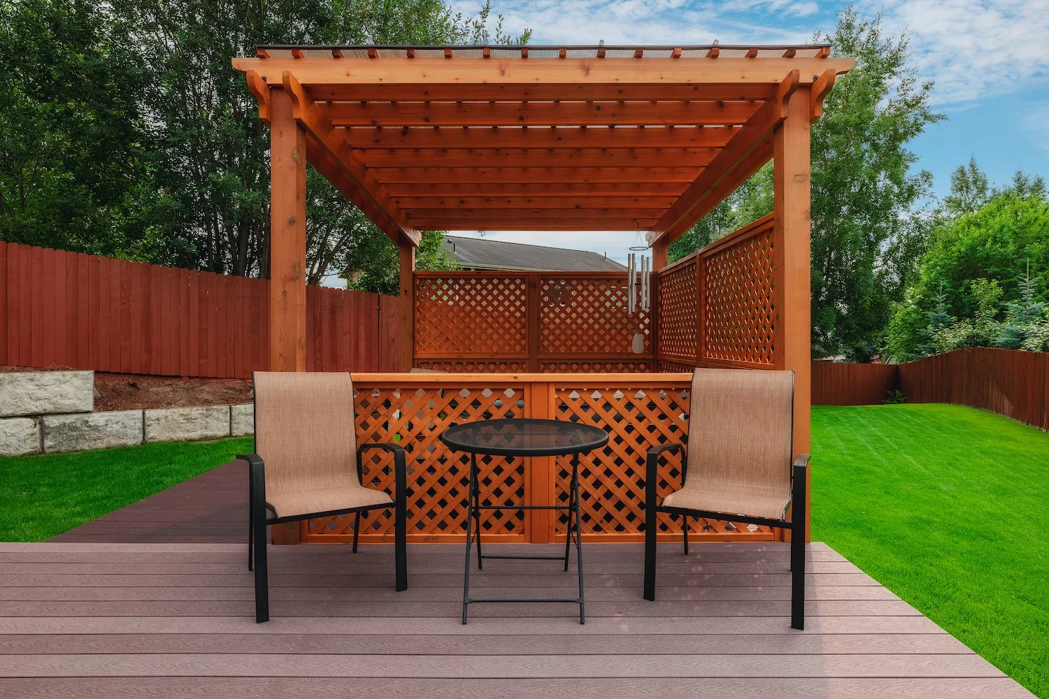 Backyard patio with two beige chairs and a small round black table on a wooden deck, with a wooden pergola and red fence, green grass, and trees.