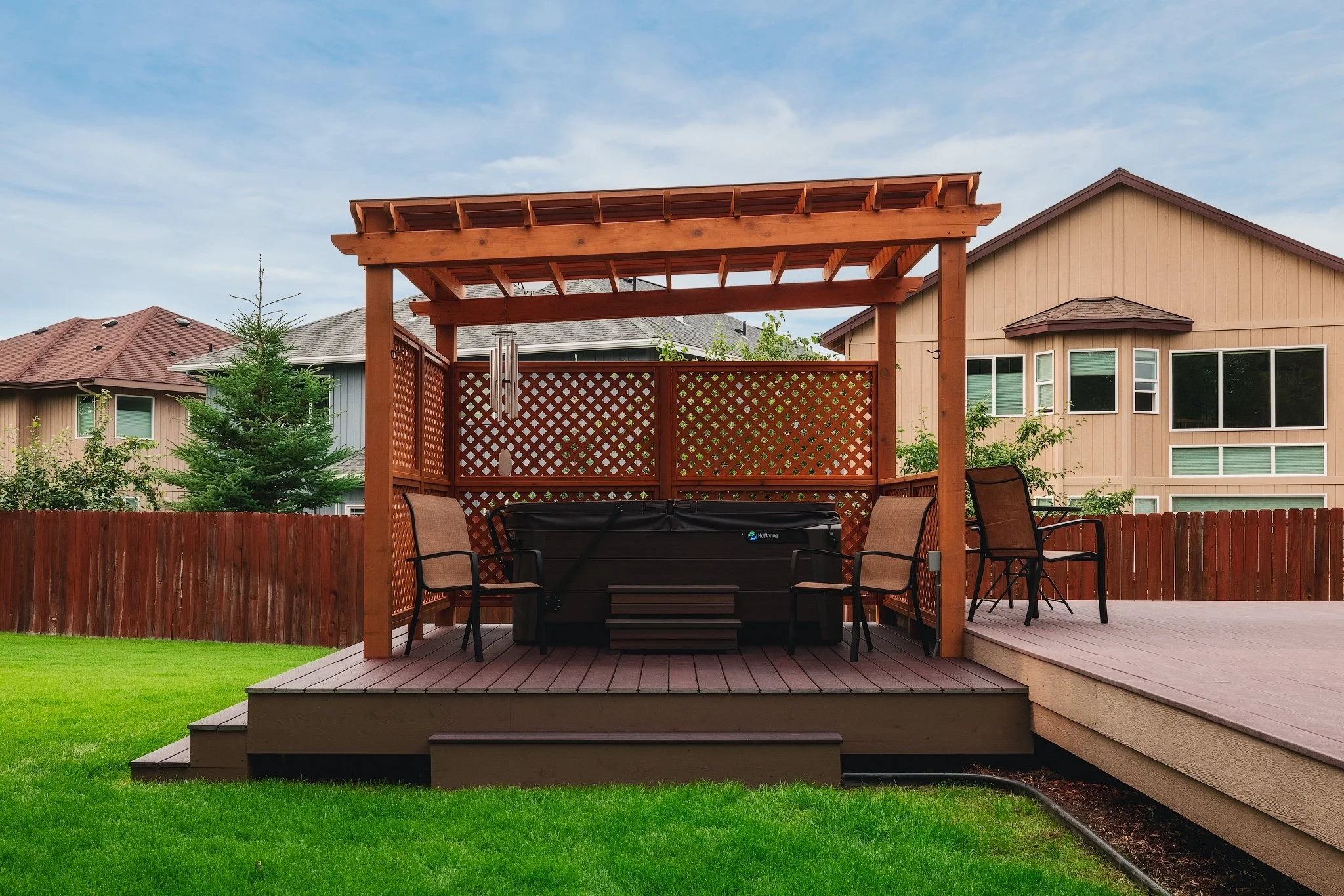 Backyard patio with a wooden pergola, hot tub, and outdoor furniture, fenced with greenery and neighboring houses in the background.