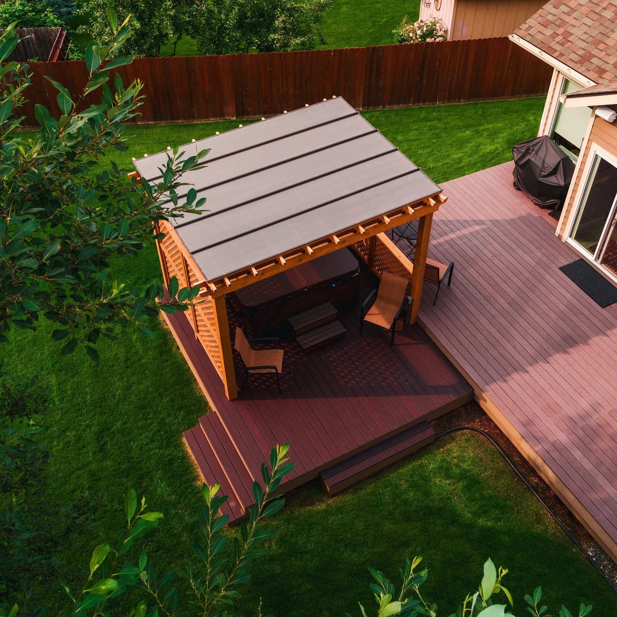 A backyard with a composite deck and a covered hot tub, outdoor chairs, and a barbecue grill. There is a small staircase leading from the deck to a grassy lawn, and a wooden privacy fence surrounds the yard.