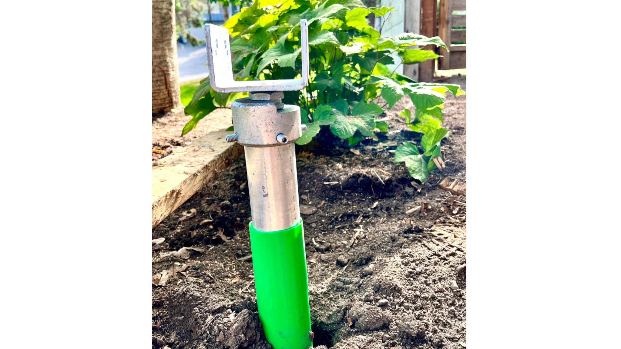A garden bed with dark soil and green plants. A metal stake with a green plastic cover at the bottom stands in the soil. The stake has an attachment at the top with an L-shaped bracket.