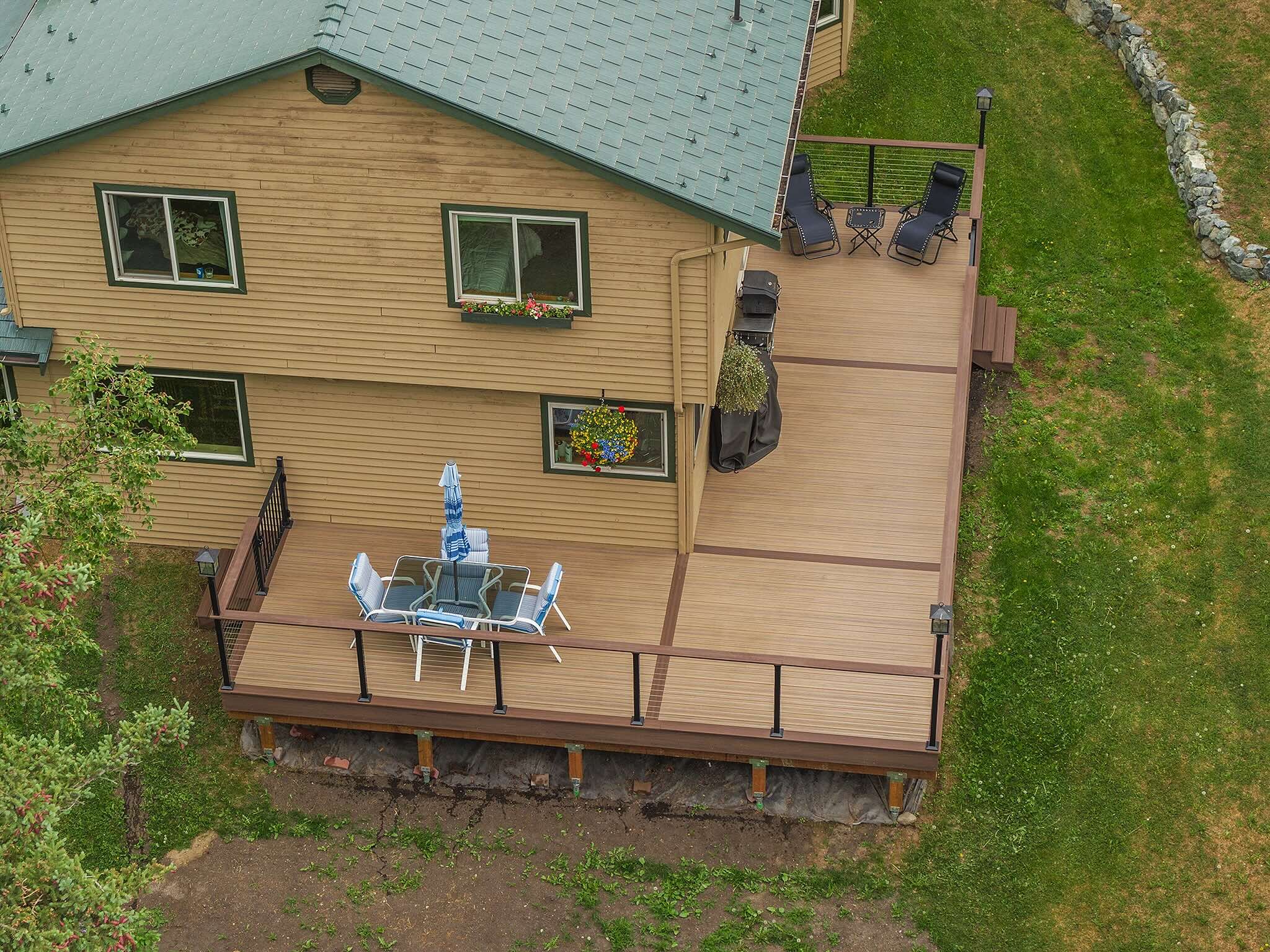 A backyard view featuring a large composite deck attached to a house with blonde siding and a gray shingled roof. The deck has seating areas with chairs, a table, and an umbrella, as well as a grill.