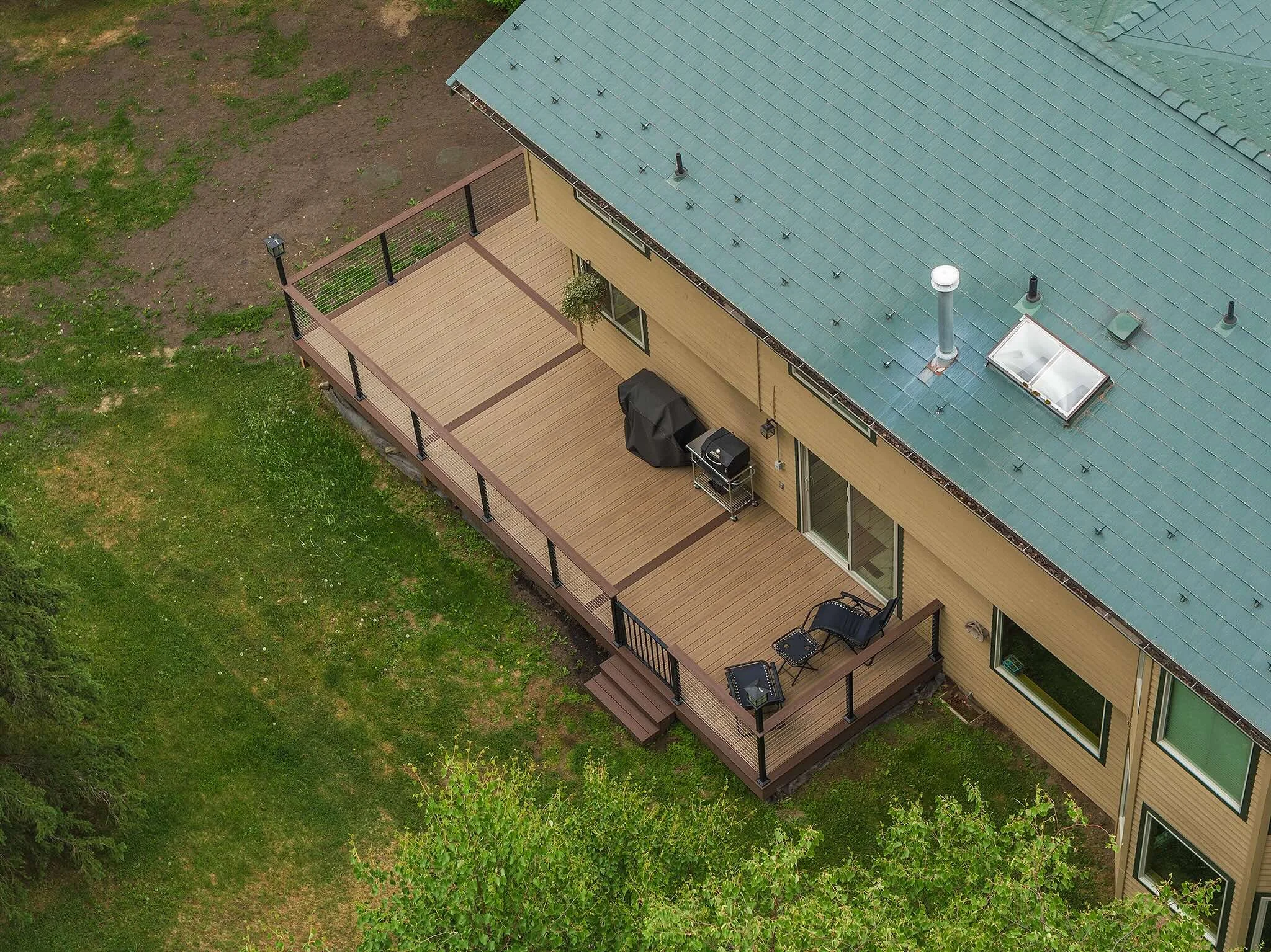 Aerial view of a house's backyard deck with outdoor chairs, a grill covered with a black cover, and a potted plant, surrounded by green grass and trees.
