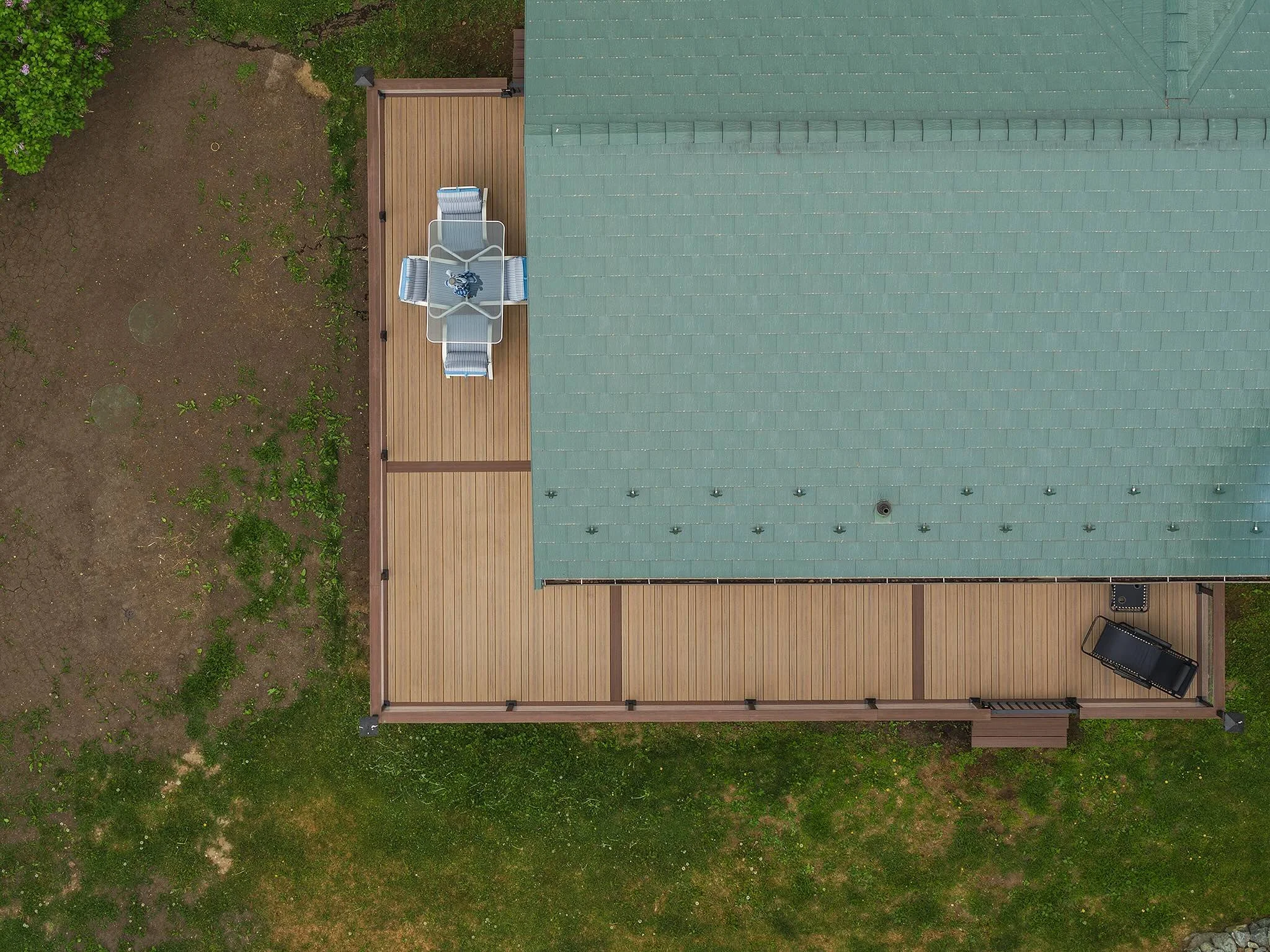 Top-down view of a house with a green shingled roof and composite deck, outdoor table with chairs, some plants and grass surrounding the house.