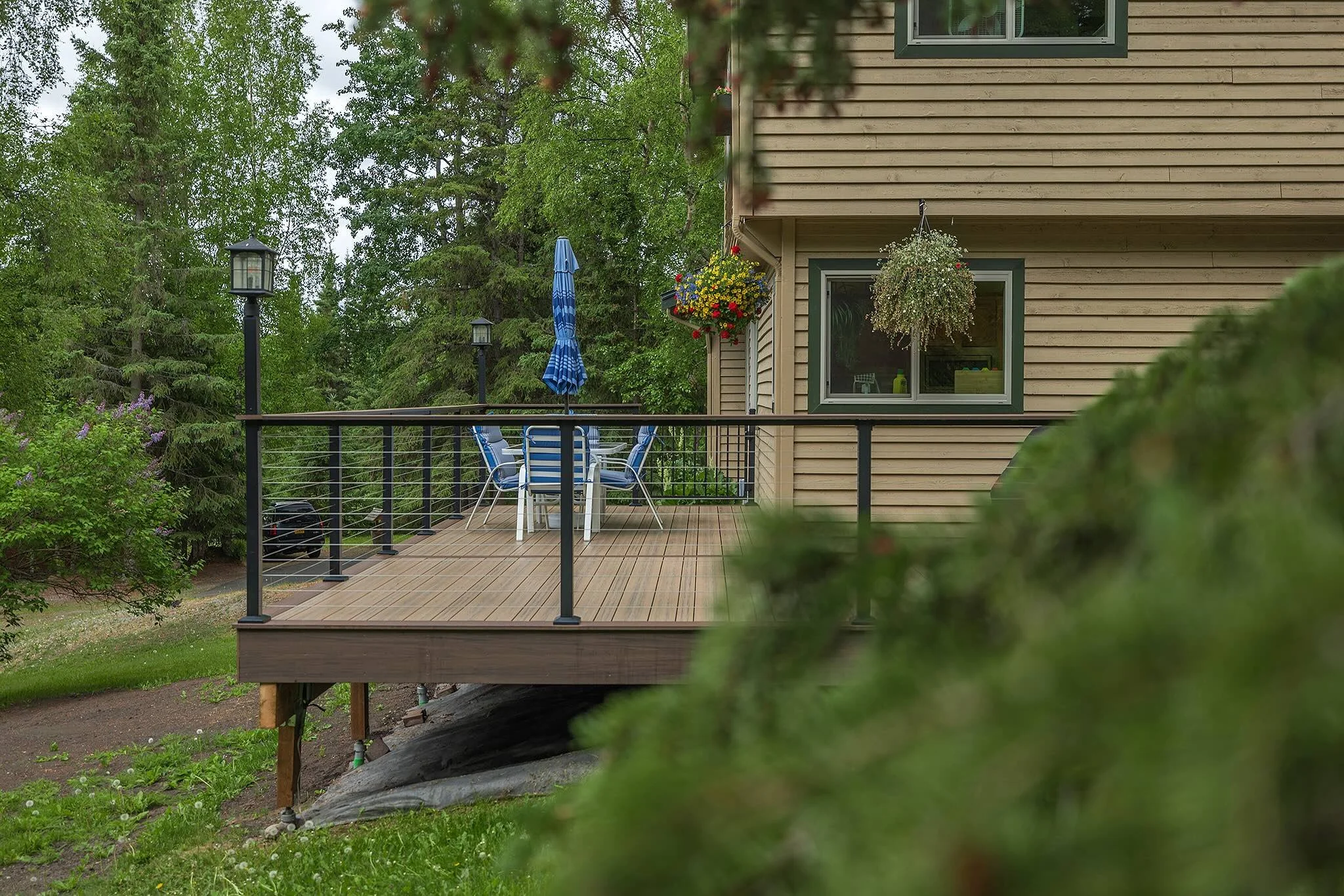 A composite deck attached to the side of a beige house with green trim, surrounded by lush green trees.  There are two lamp posts along the deck railing.
