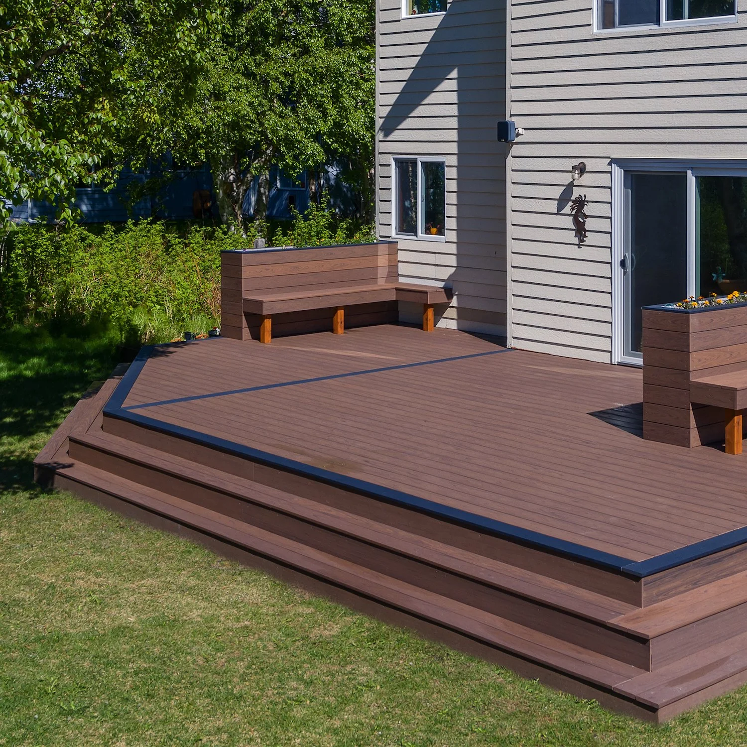 A composite deck attached to a house with stairs leading down to a grassy yard. The deck has built-in benches and flower planters, with a tree and bushes in the background.